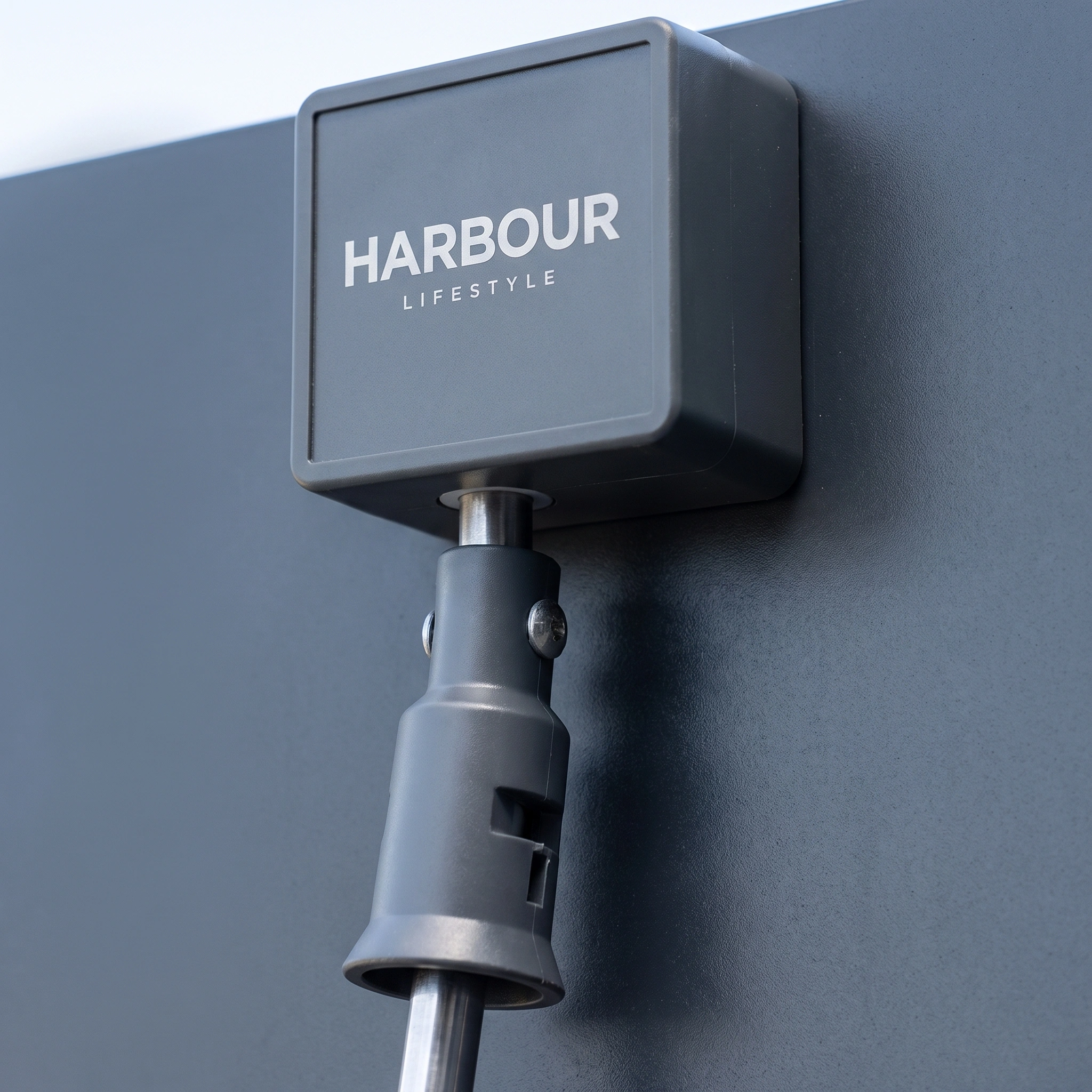 Close-up of a grey box labeled "Harbour Lifestyle" attached to a metal pole against a grey background, evoking innovative outdoor features that may be included in the PergoSTET Lite 2.4m x 3m Metal Rectangular Pergola in Grey, such as tilting louvres or an integrated drainage system for enhanced modern design.