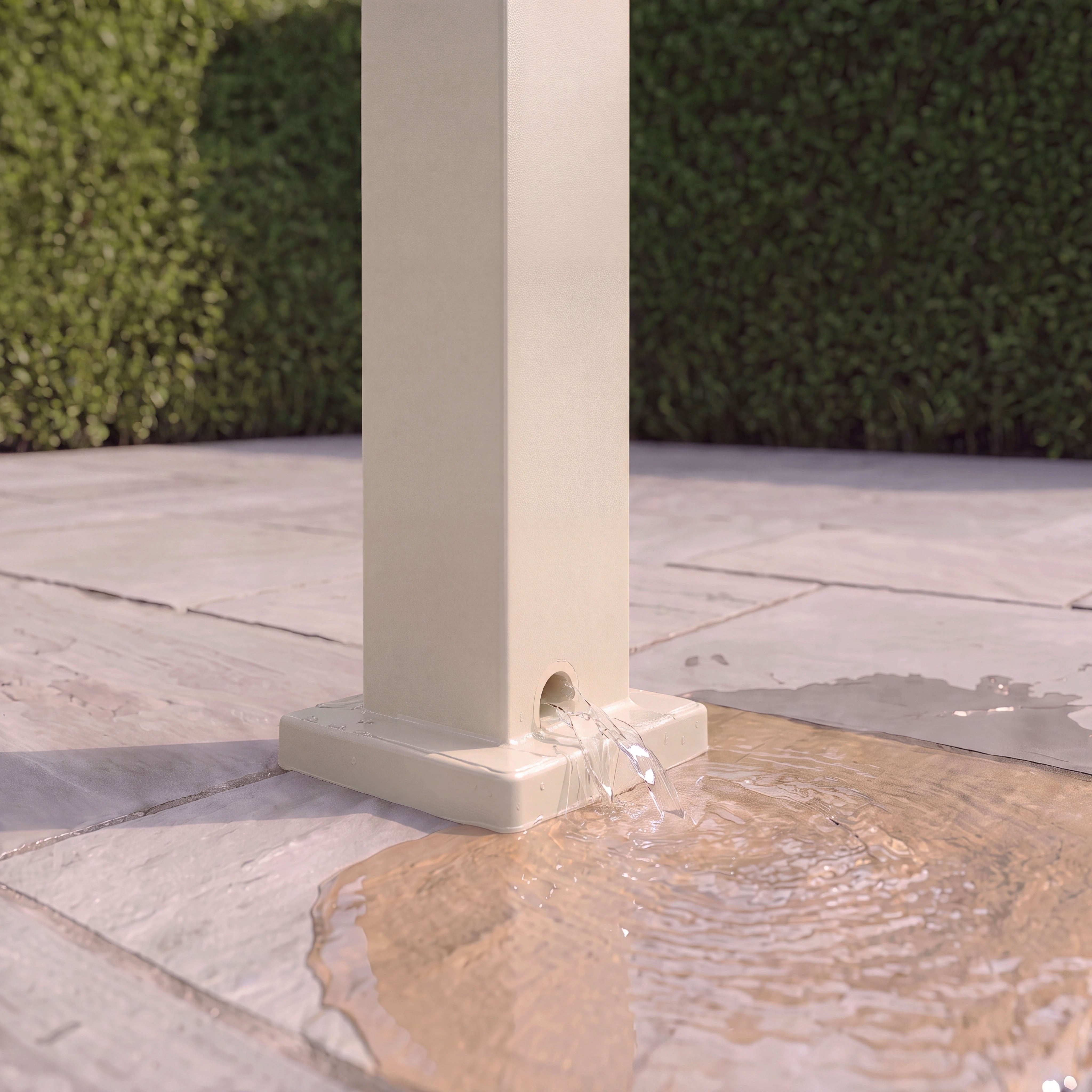 Water flows from a hole near the base of a square beige post onto stone pavement, creating a tranquil scene beneath the PergoSTET Lite 2.4m x 3m Metal Rectangular Pergola in Latte, which features tilting louvres for adjustable outdoor shelter.