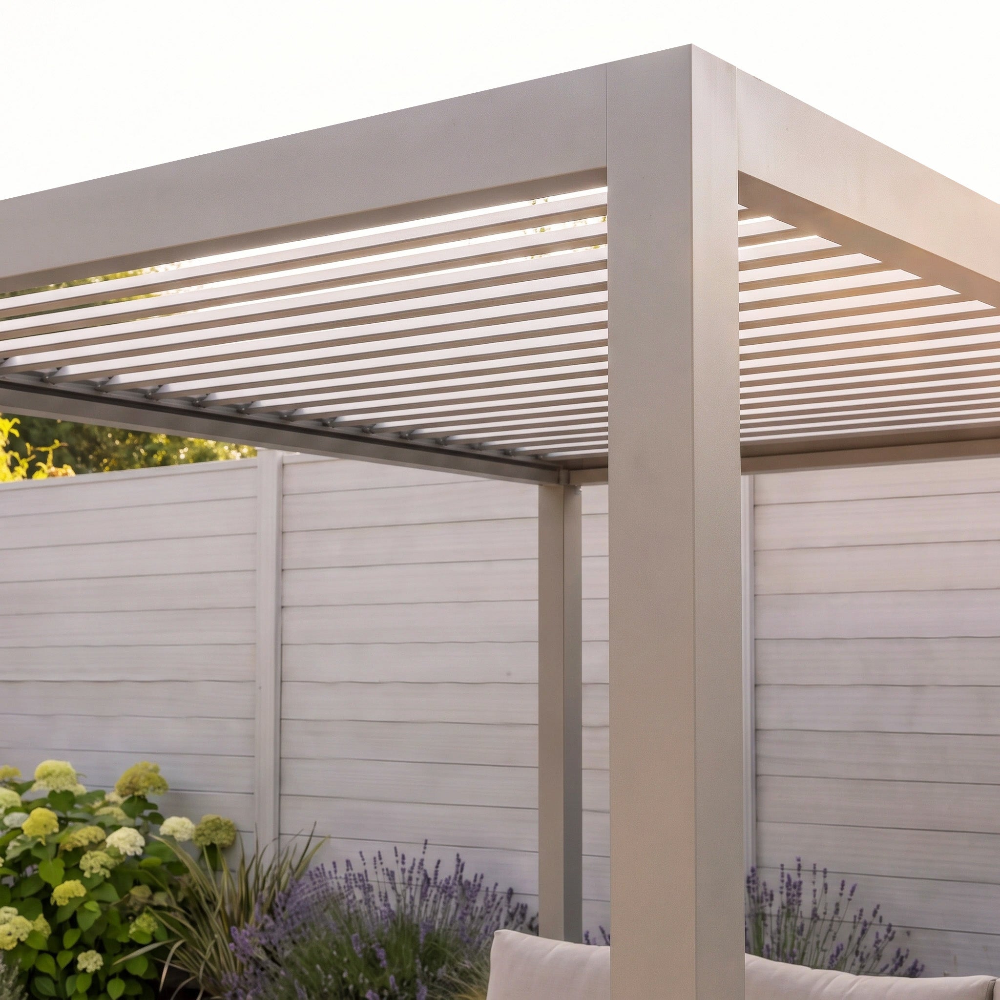 The PergoSTET Lite 2.4m x 3m Metal Rectangular Pergola in Latte features horizontal slats and tilting louvres, creating an elegant outdoor shelter in a garden setting near a white fence and lush green plants at sunset.