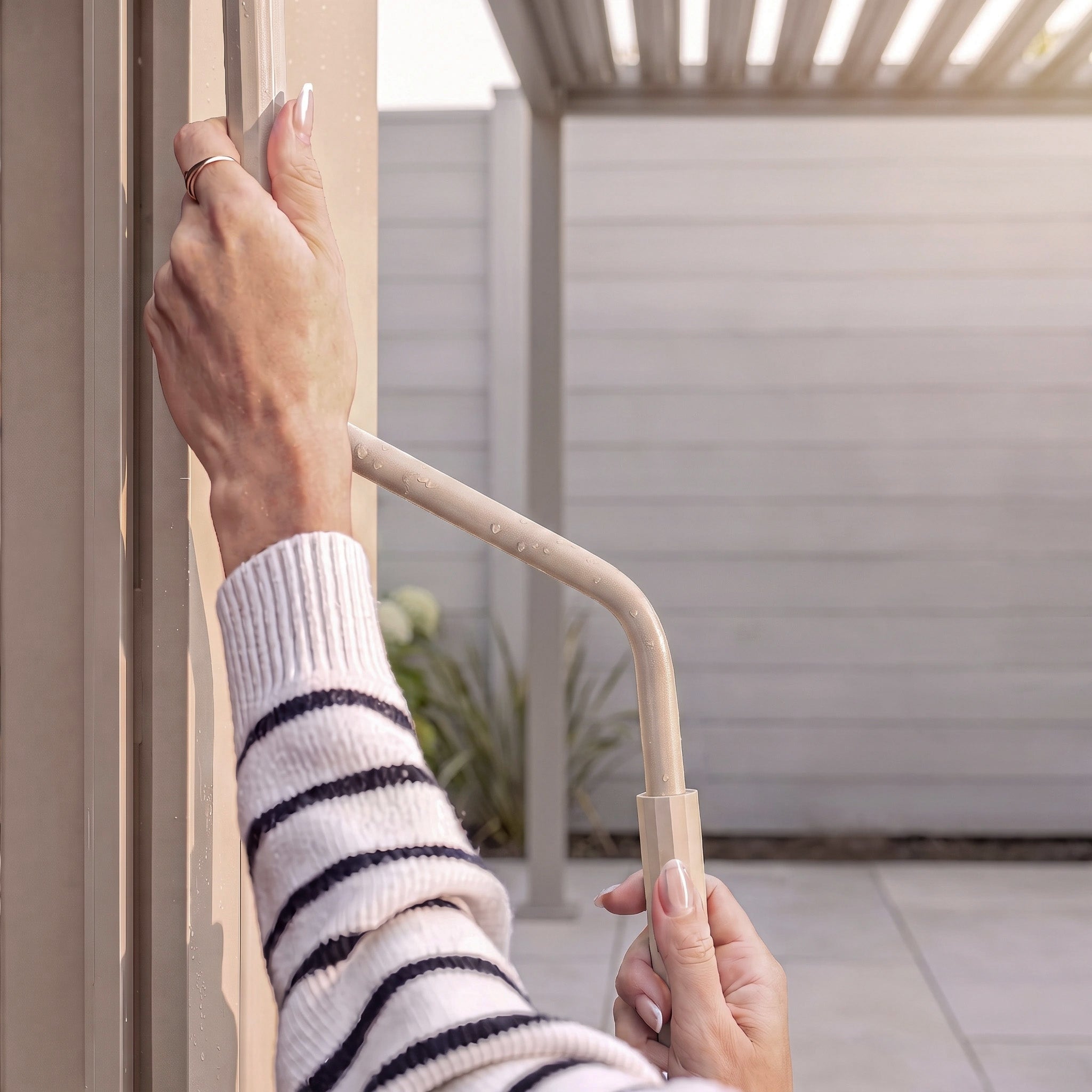 Hands turning a beige crank handle on the PergoSTET Lite 2.4m x 3m Metal Rectangular Pergola in Latte, featuring tilting louvres; a person in a striped sweater enjoys the comfort of this stylish outdoor shelter.