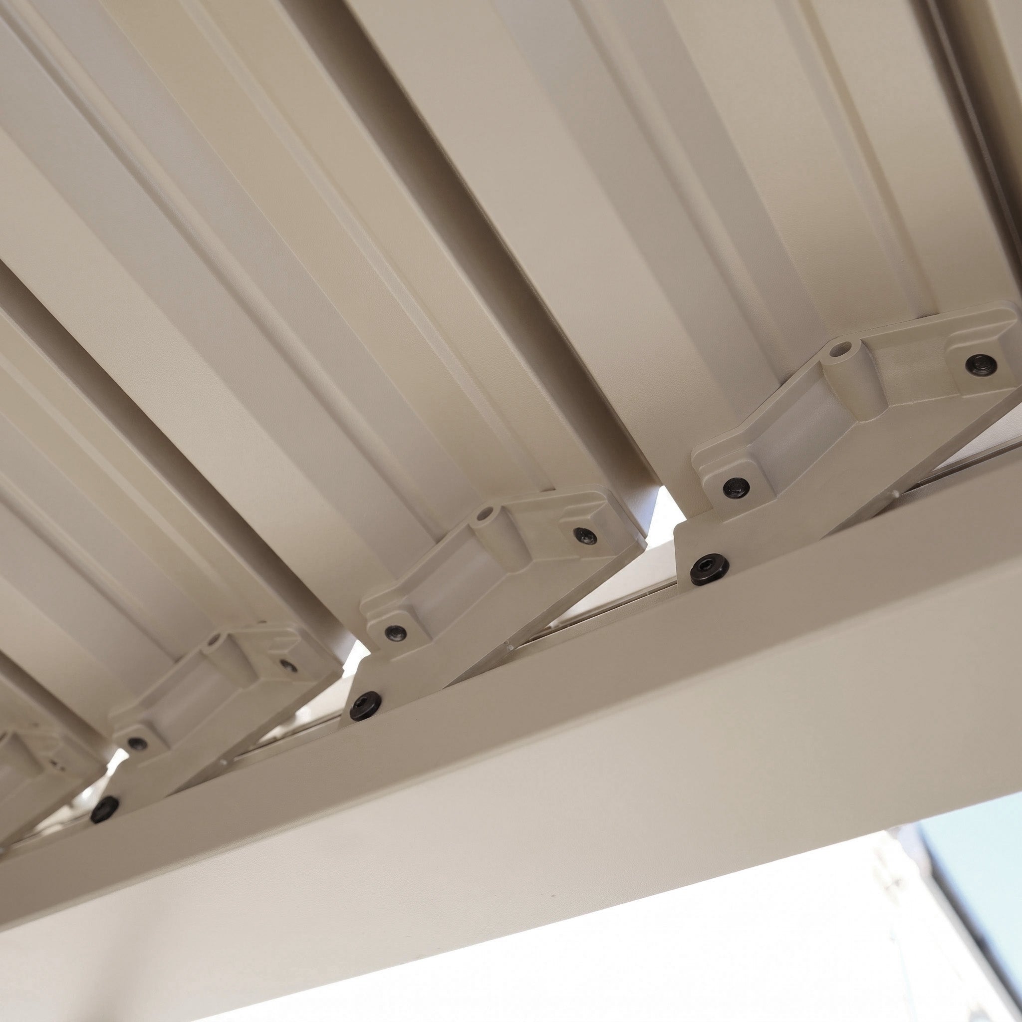 Close-up view of the PergoSTET Lite 2.4m x 3m Metal Rectangular Pergola in Latte, highlighting its beige louvered roof from beneath with visible screws and joints, characteristic of stylish outdoor shelters with tilting metal louvres.