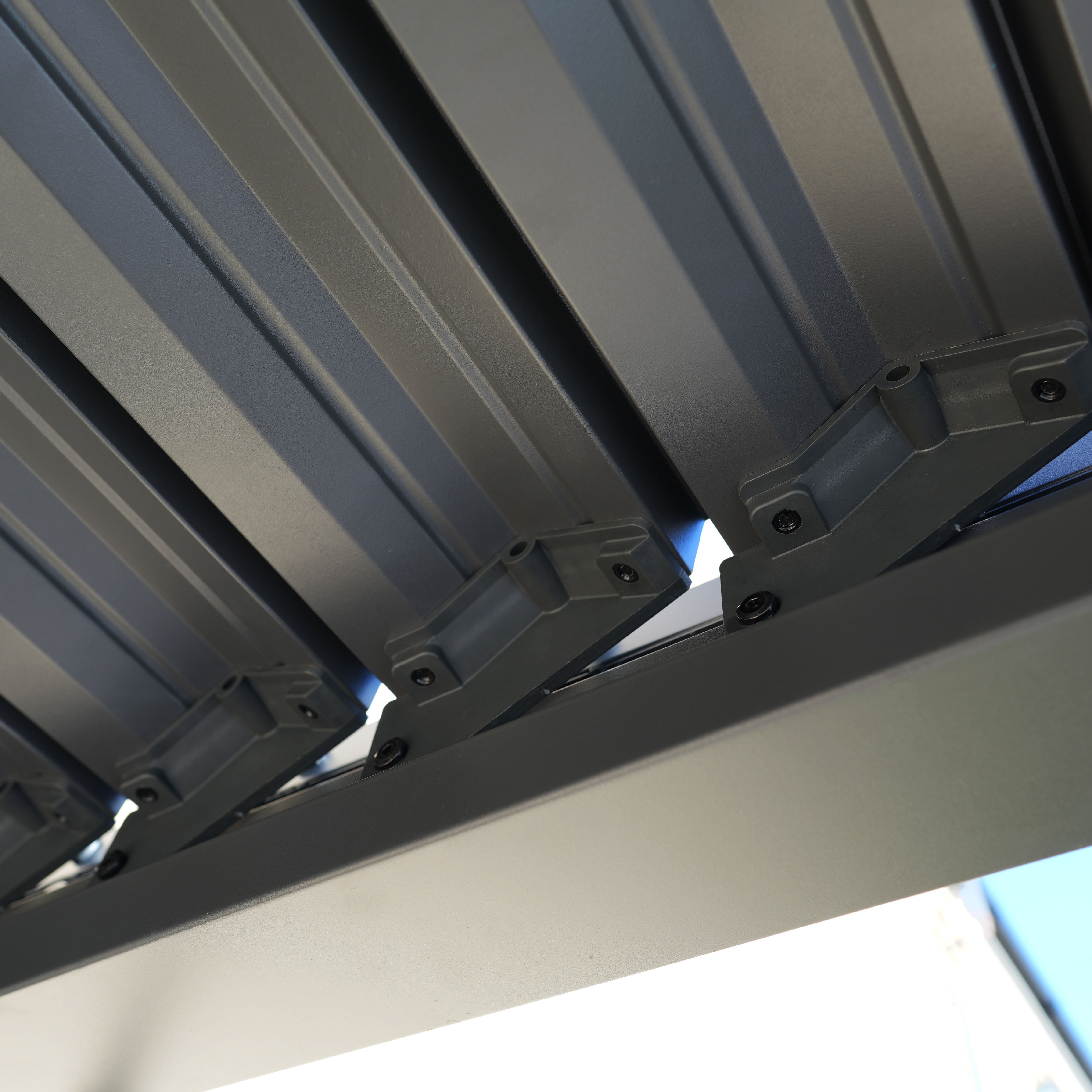 Close-up of metal beams and brackets on the PergoSTET Lite 2.4m x 3m Metal Rectangular Pergola in Grey, featuring tilting louvres, viewed from below against the sky.