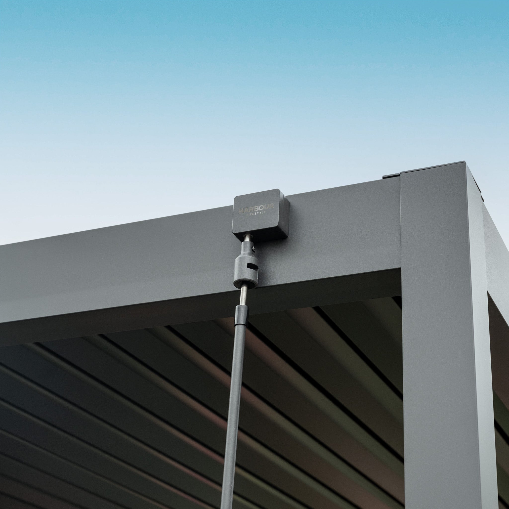 Close-up of a black device attached to the corner of a modern grey PergoSTET Lite Neo 3m x 7m Metal Rectangular Pergola with tilting steel louvres, set against a clear blue sky.
