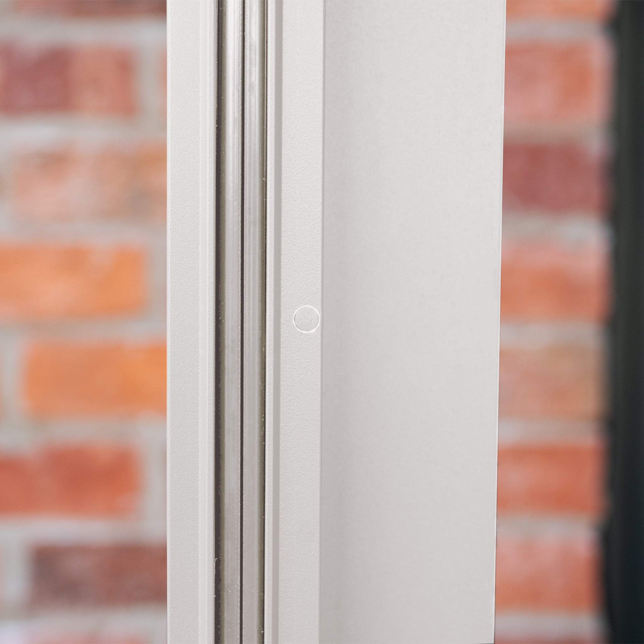 Close-up of a white metal frame with a small circular mark, set against a blurred brick wall—an elegant detail of the PergoSTET Plus 3m x 4m Rectangular Pergola with 3 Drop Sides and LED Lighting in Latte, featuring a built-in drainage system.