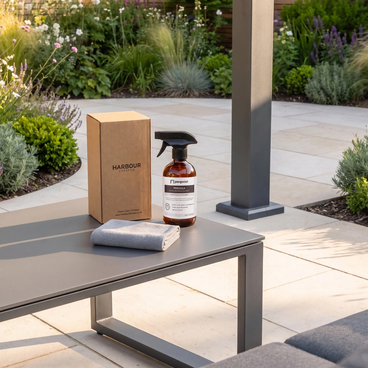 Outdoor Metal Pergola Cleaner, along with a box and cloth, sits on a patio table in a lush garden filled with flowers and plants—perfect essentials for deep cleaning your metal pergola.