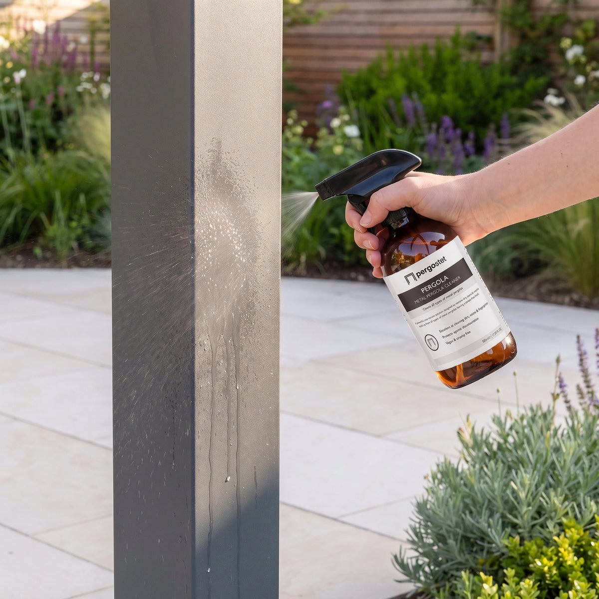 A hand sprays Outdoor Metal Pergola Cleaner onto a gray outdoor post in a landscaped garden with stone tiles.