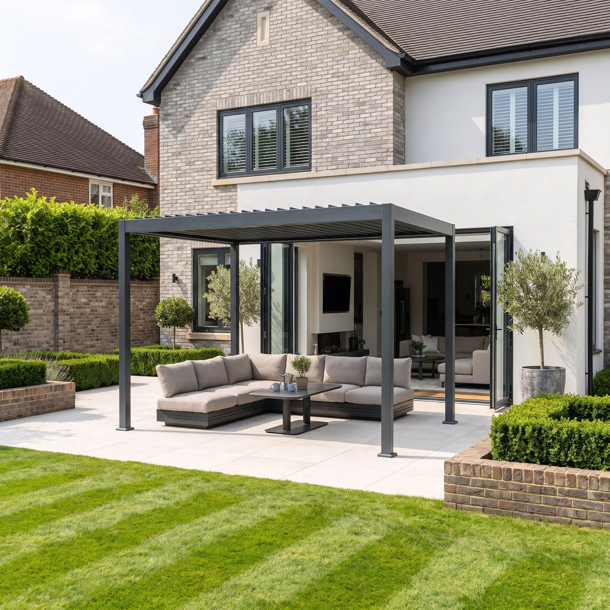 Modern house patio with a PergoSTET Lite 3m x 4m Metal Rectangular Pergola featuring motorised louvres and integrated LED lighting in grey, complemented by outdoor sofas, coffee table, and manicured lawn on a sunny day.