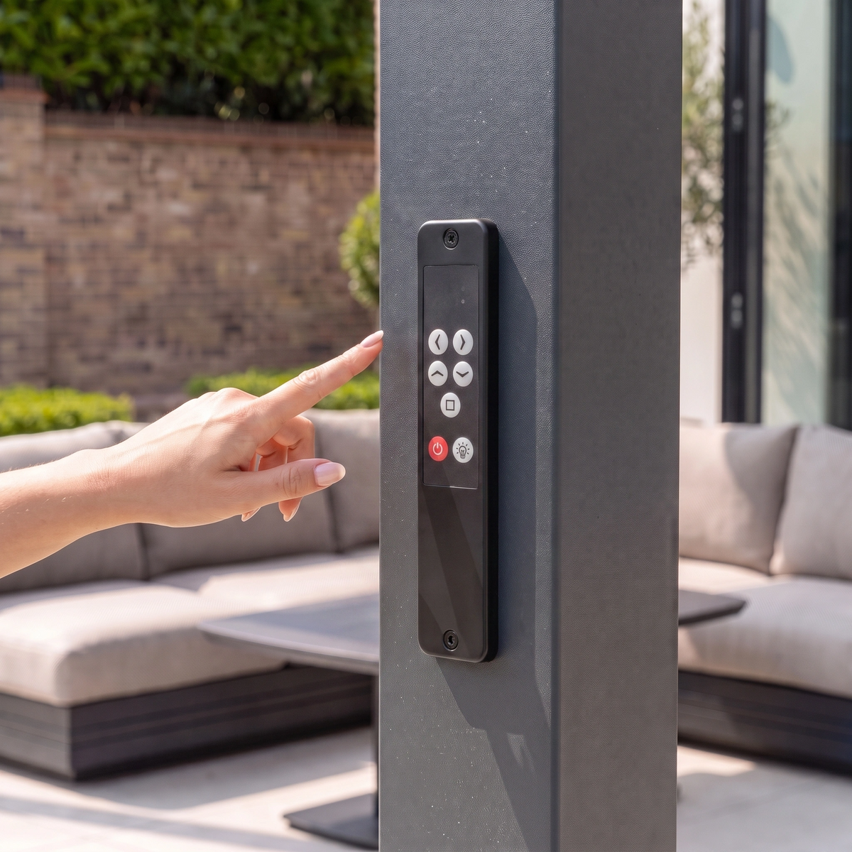 A hand presses a button on an outdoor control panel near patio furniture, with plants in the background and the sleek PergoSTET Lite 3m x 4m Metal Rectangular Pergola With Motorised Louvres and LED Lighting in Grey overhead.