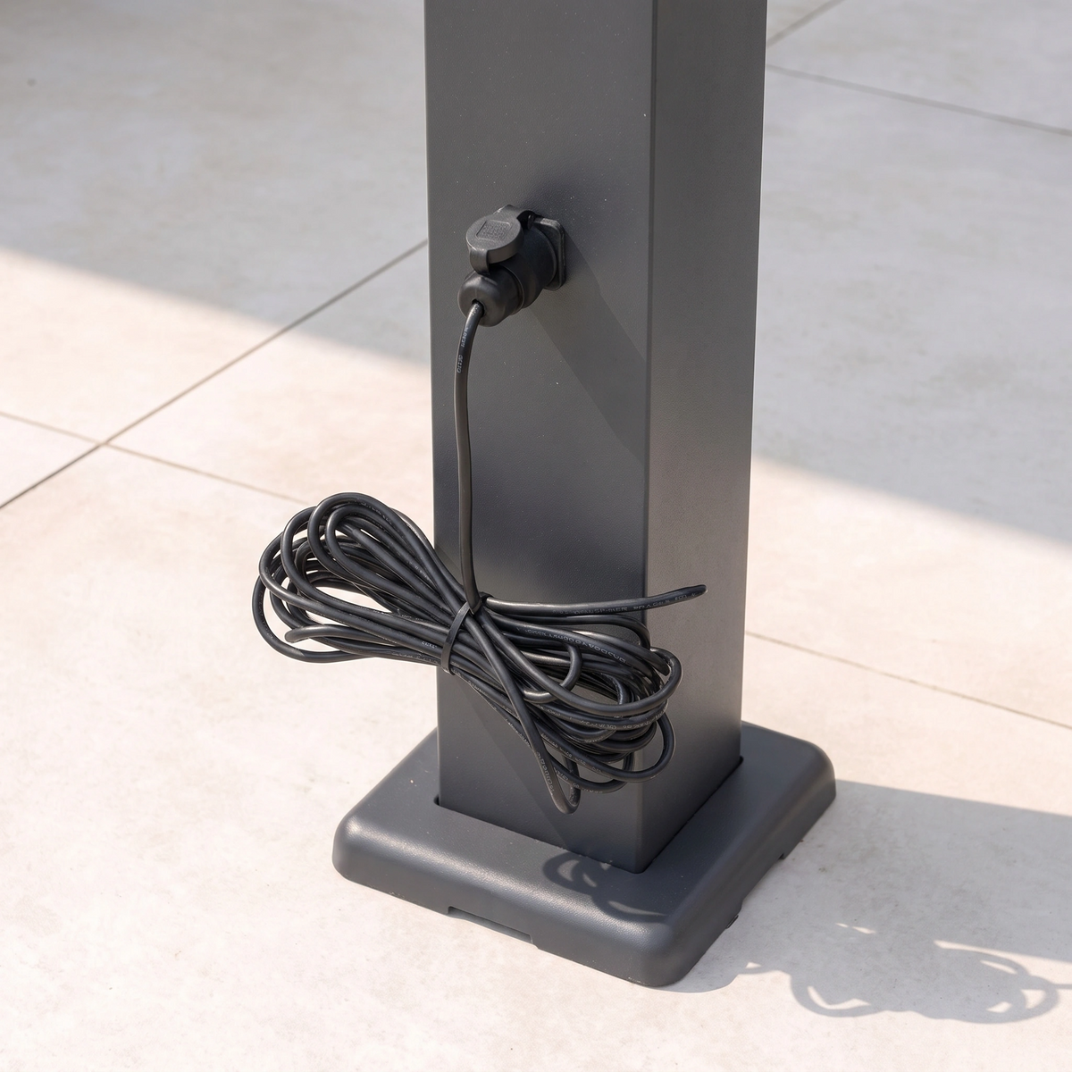 A black electric cable is plugged into an outdoor socket on a grey metal post, coiled neatly at the base, ready to power features such as LED lighting or motorised louvres for the PergoSTET Lite 3m x 4m Metal Rectangular Pergola With Motorised Louvres and LED Lighting in Grey.
