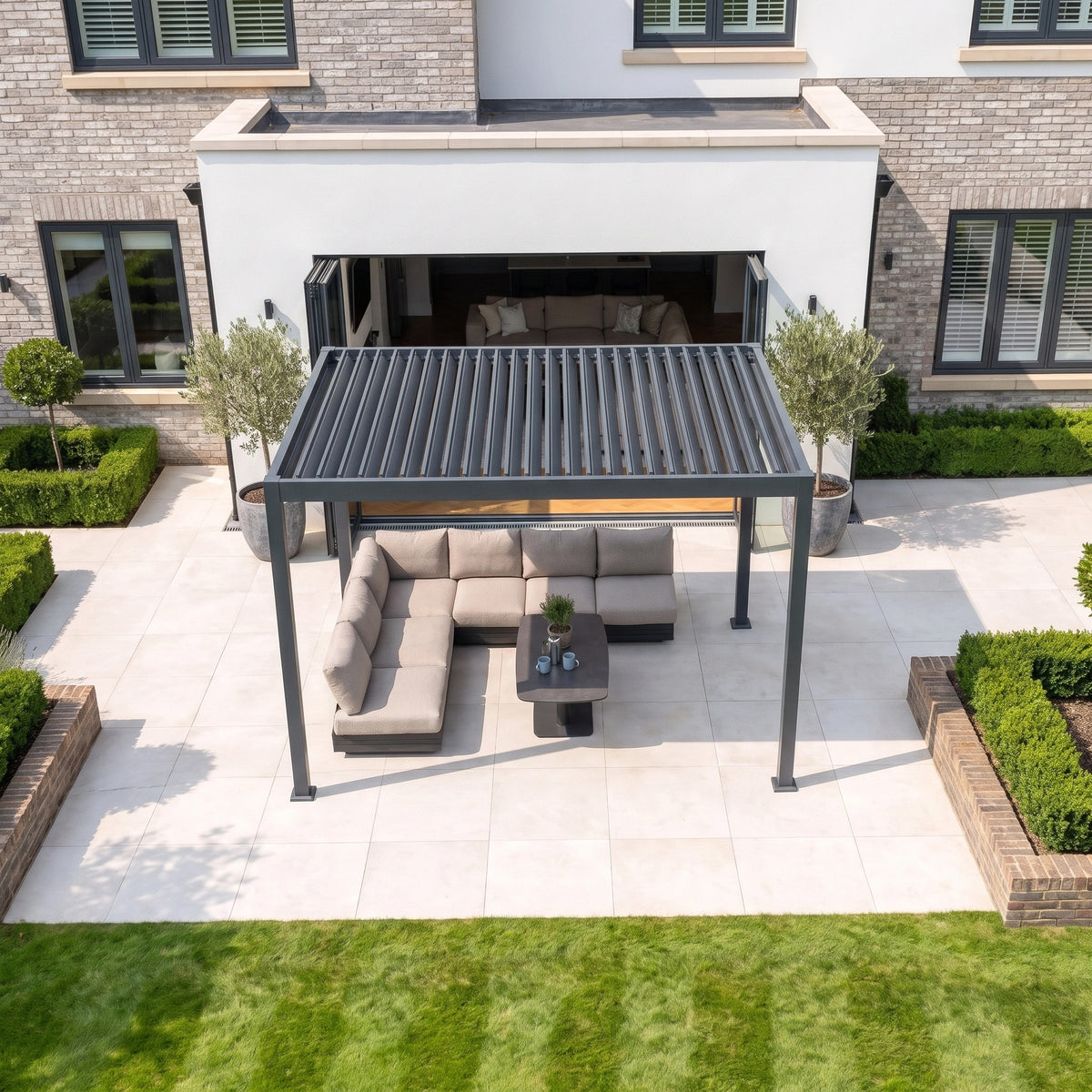 A PergoSTET Lite 3m x 4m Metal Rectangular Pergola with Motorised Louvres and LED Lighting in Grey covers a sectional outdoor sofa and table on a patio beside a landscaped garden.