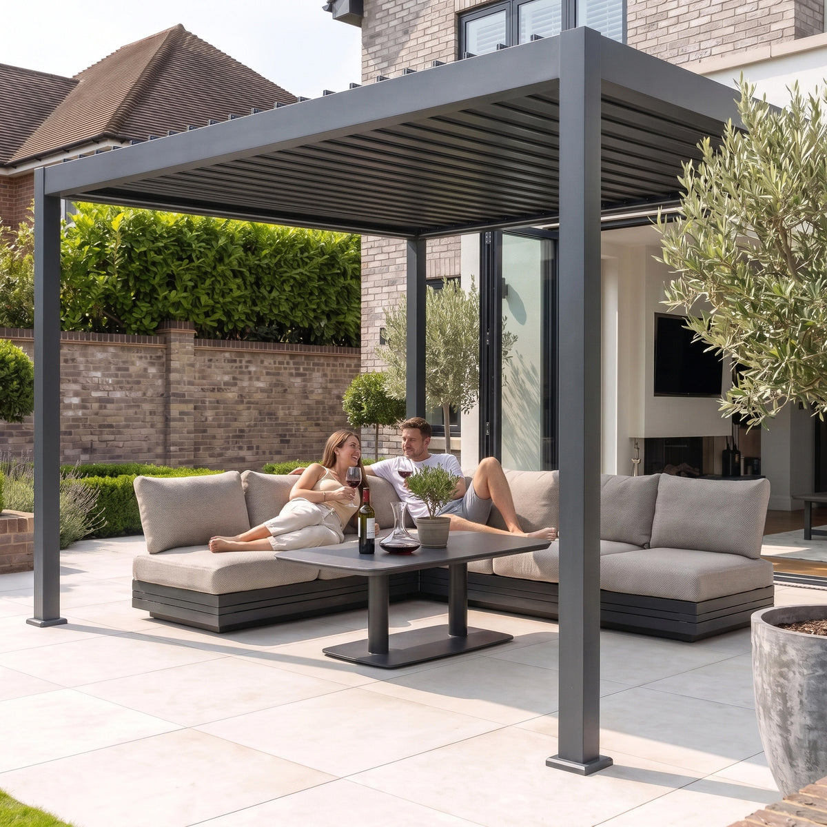 A couple relaxes on a patio sofa beneath the PergoSTET Lite 3m x 4m Metal Rectangular Pergola With Motorised Louvres and LED Lighting in Grey, sharing wine in their sunny backyard.
