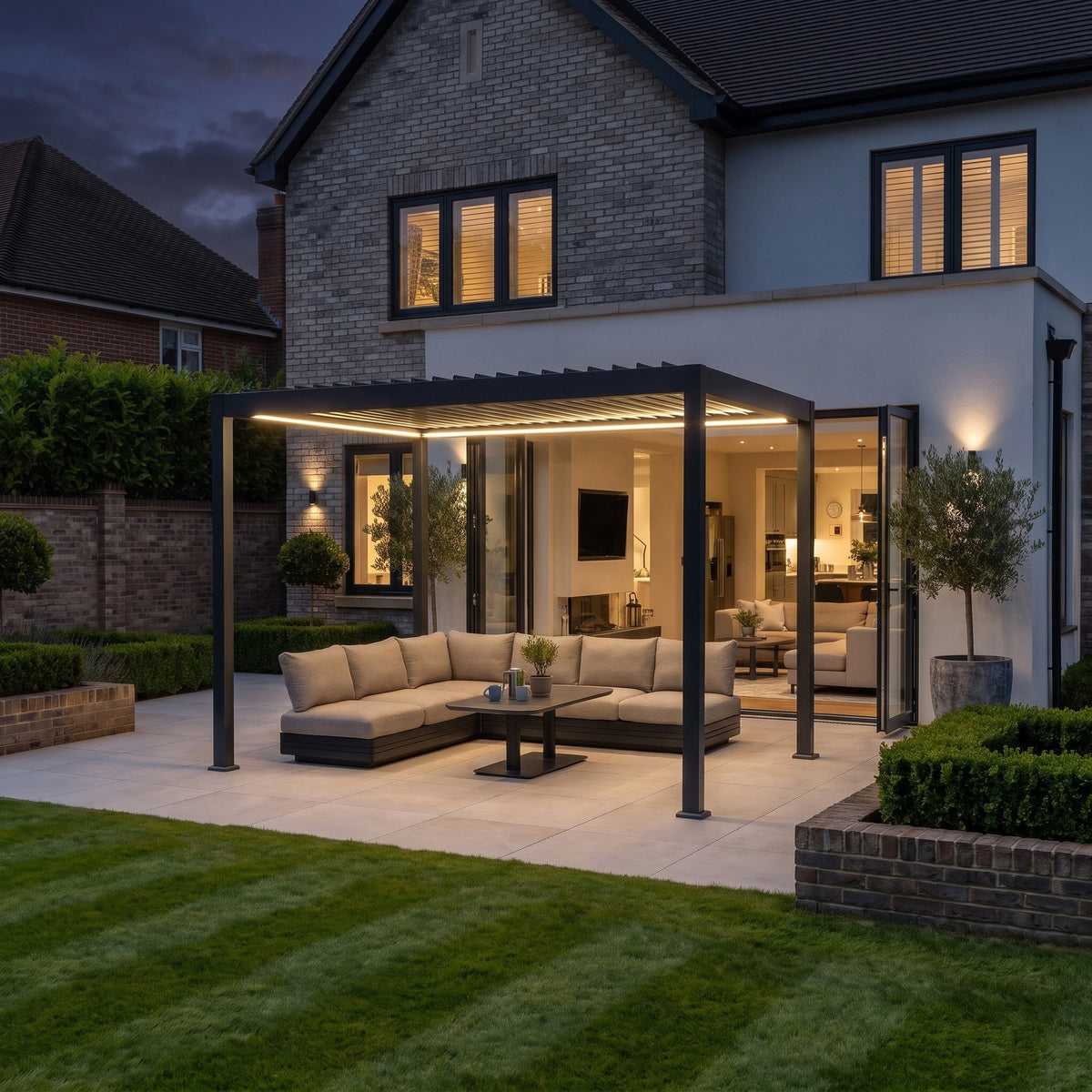 A modern patio features the PergoSTET Lite 3m x 4m Metal Rectangular Pergola in Grey, equipped with motorised louvres and LED lighting, sheltering an outdoor sofa set and adjoining a contemporary house at dusk, with soft illumination highlighting the inviting interior.