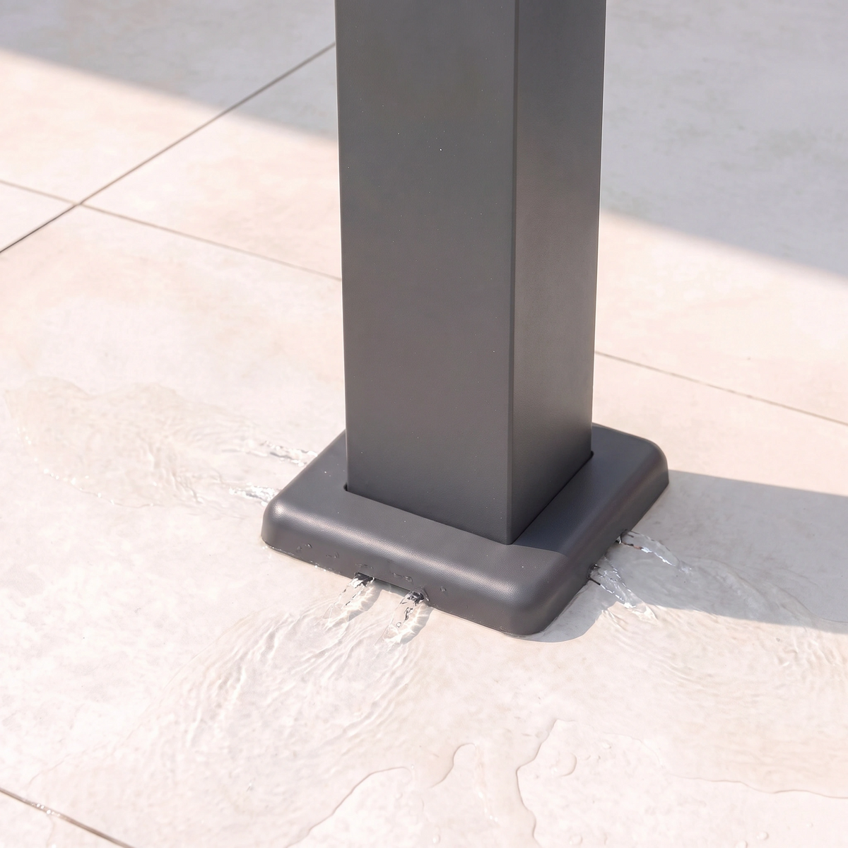 A black metal post base with water pooling around it on light-colored tiled flooring in sunlight, adjacent to the PergoSTET Lite 3m x 4m Metal Rectangular Pergola With Motorised Louvres and LED Lighting in Grey.