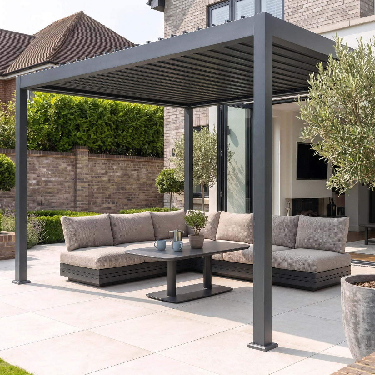 A modern outdoor patio showcases the PergoSTET Lite 3m x 4m Metal Rectangular Pergola With Motorised Louvres and LED Lighting in Grey, complemented by cushioned sofas, a round table, and lush backyard greenery.
