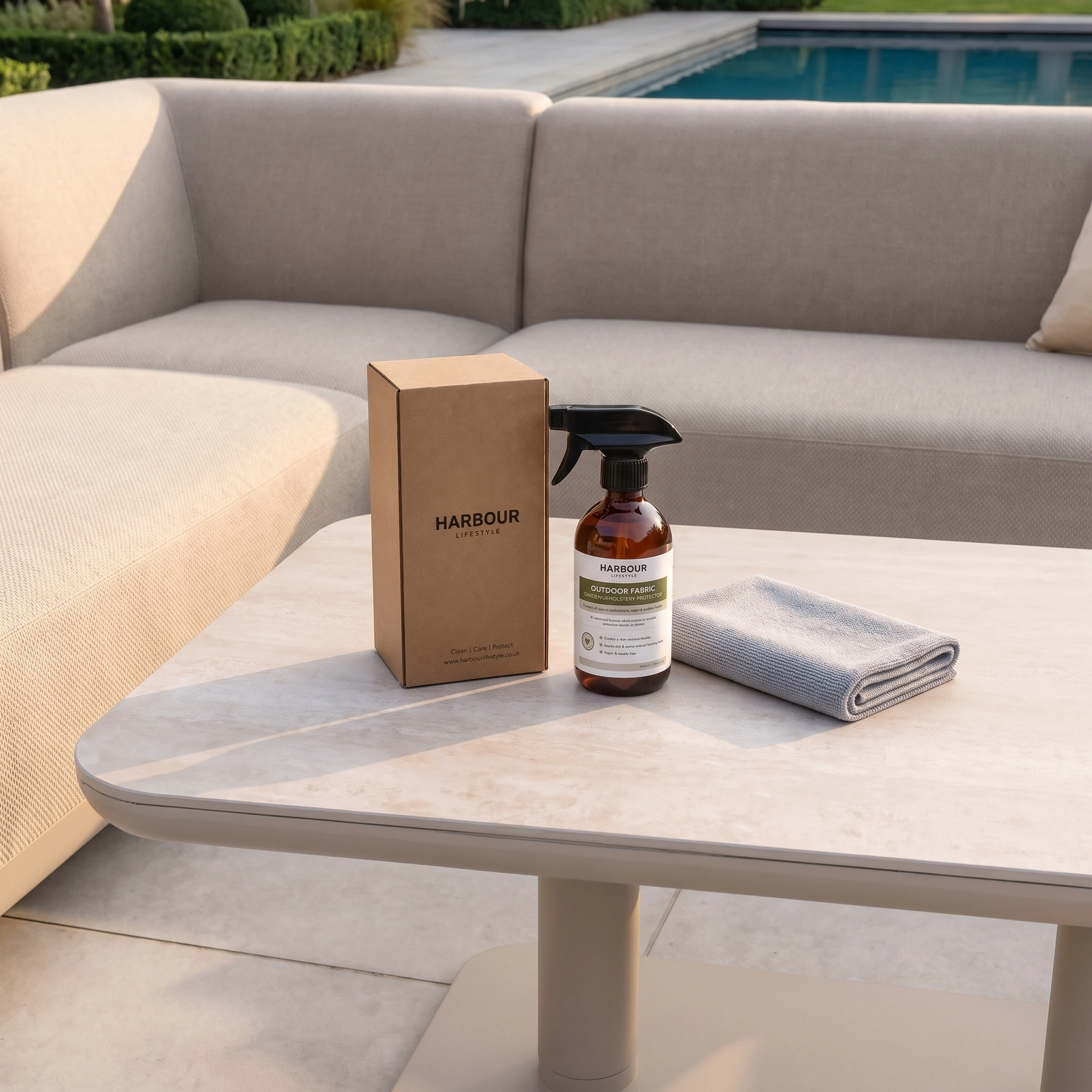 A spray bottle of Outdoor Furniture Cushion, Rope and Upholstery Protector, a box, and a cloth sit on an outdoor table near beige seating and a pool.