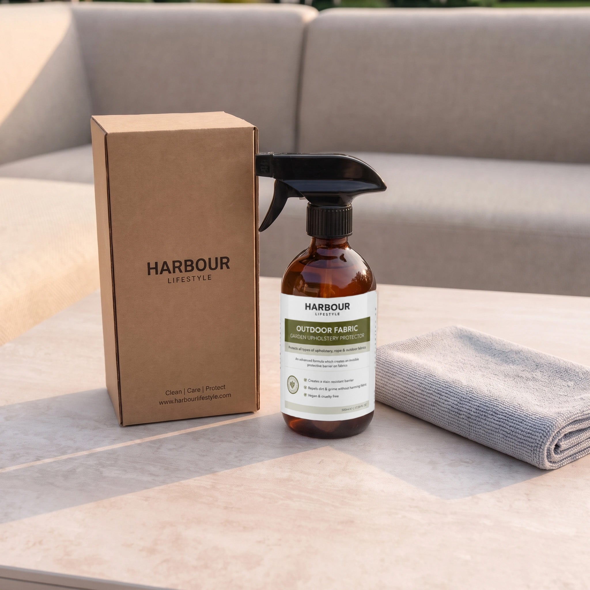 Bottle of outdoor fabric cleaner, box, and gray cloth on a table with a couch in the background—perfect for pairing with your favorite Outdoor Furniture Cushion, Rope and Upholstery Protector to keep cushions fresh and long-lasting.