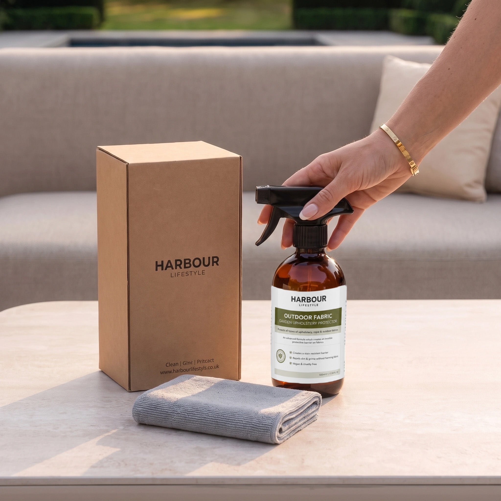 A hand holds a spray bottle next to a box labeled “Harbour” on a table, with a gray cloth and outdoor sofa—perfect for applying the Outdoor Furniture Cushion, Rope and Upholstery Protector to keep your cushions looking fresh and clean.