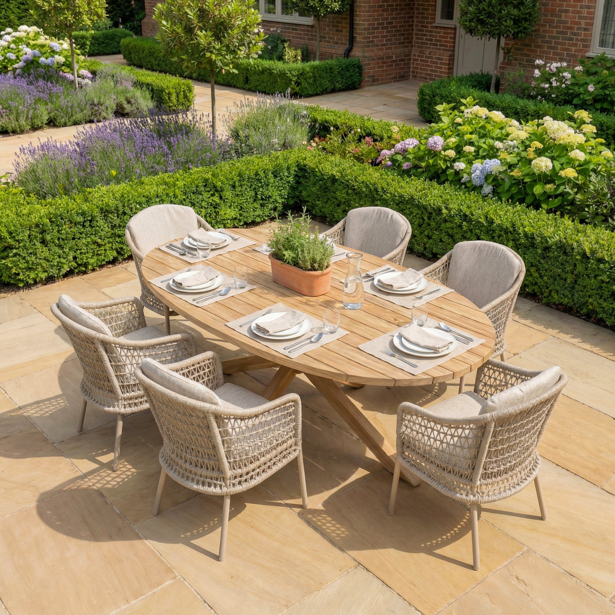 The Puccini 6 Seat Oval Dining Set in Latte features a wood patio table with six cushioned chairs, ideal for dining and beautifully set amid garden plants and flowers.