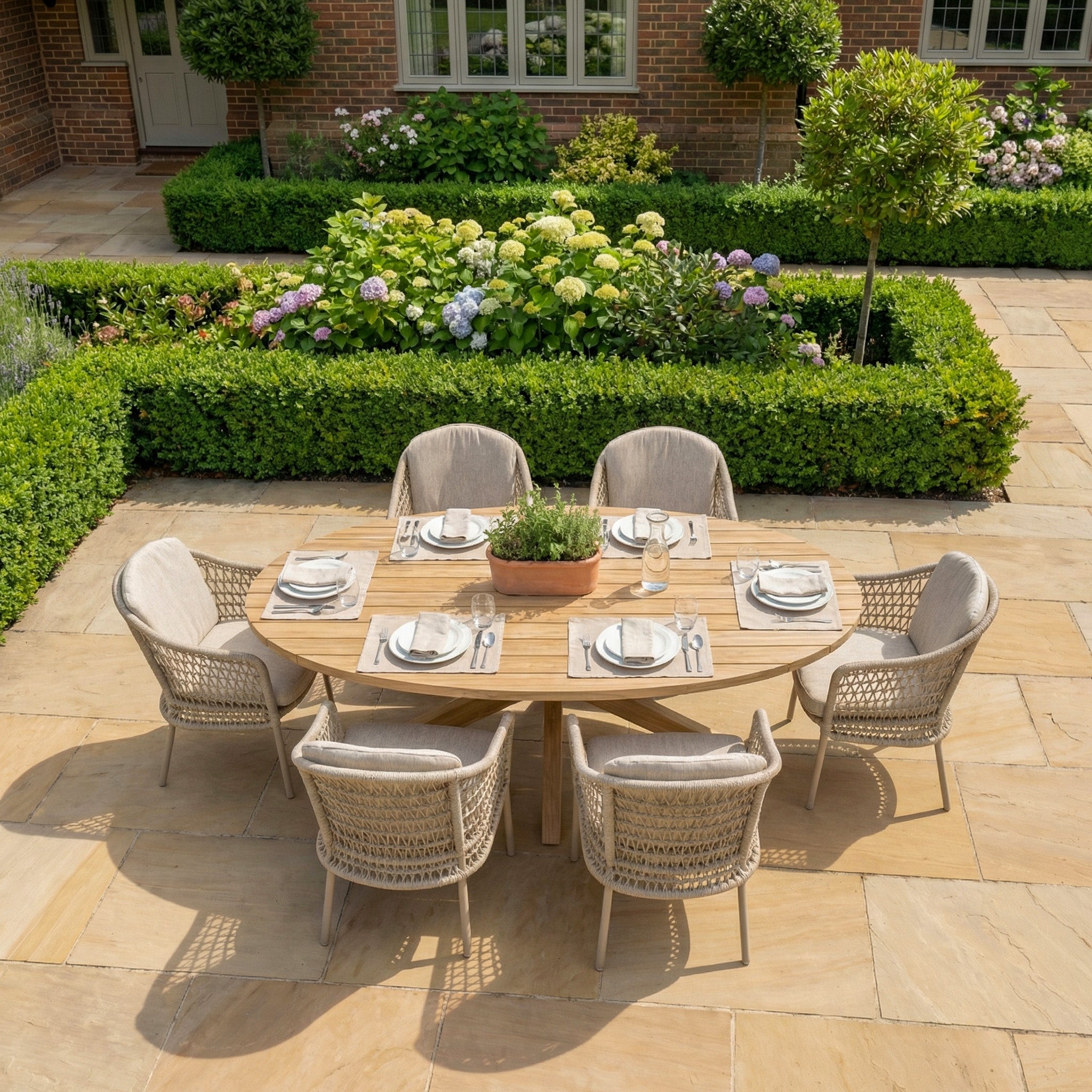 The Puccini 6 Seat Oval Dining Set in Latte, arranged on a stone patio and surrounded by garden flowers, offers a stylish and inviting addition to your outdoor space.