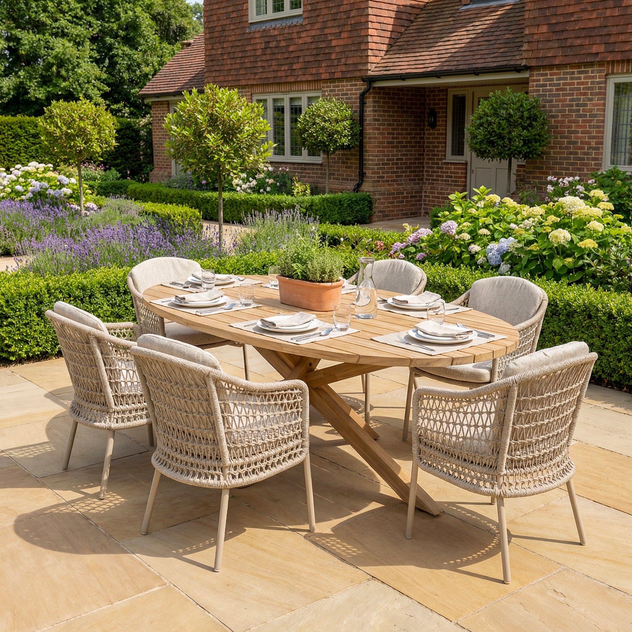 The Puccini 6 Seat Oval Dining Set in Latte is arranged on a garden patio near a brick house.