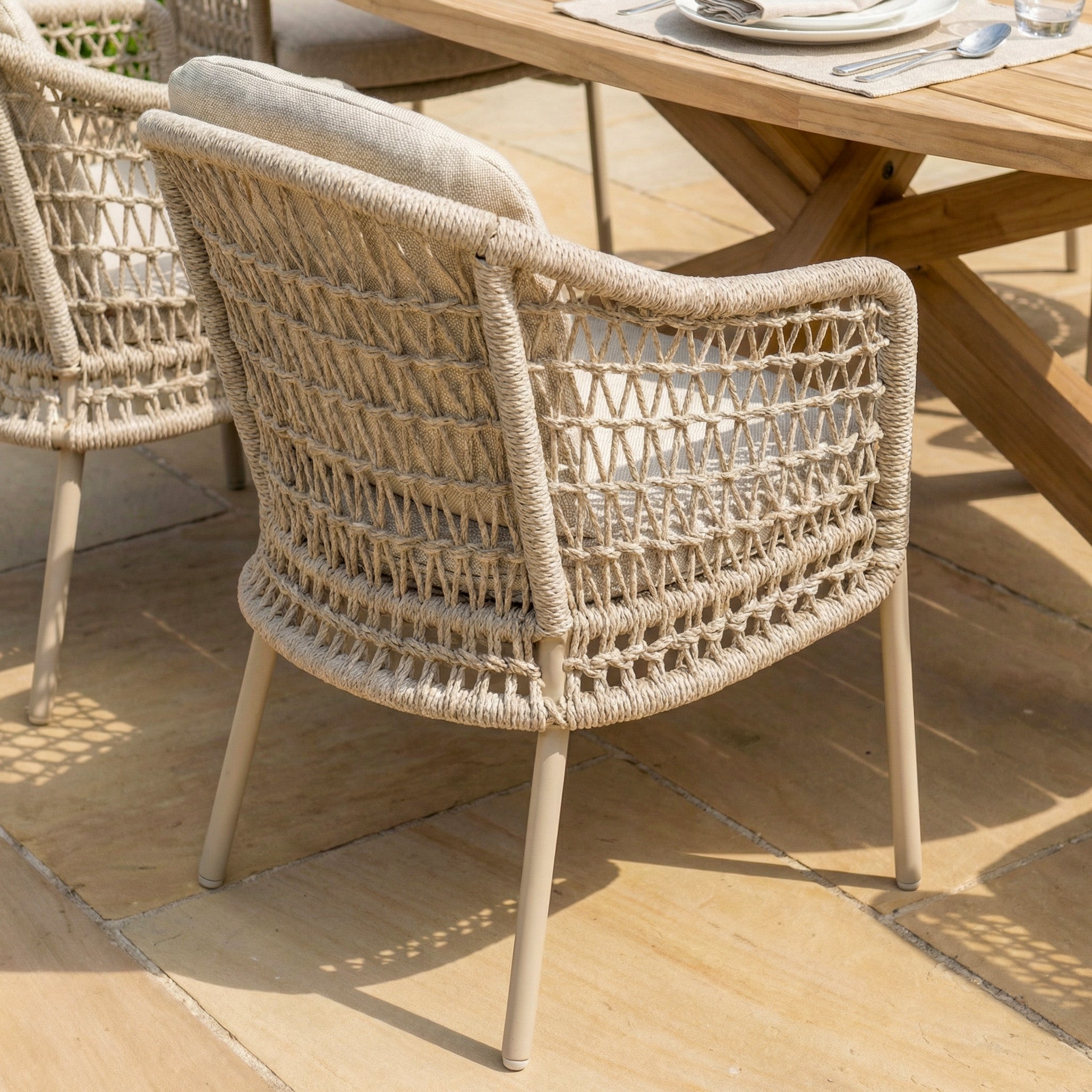 The Puccini 6 Seat Oval Dining Set in Latte, featuring woven rattan chairs with metal legs, is arranged beside a wooden dining table on stone patio tiles.