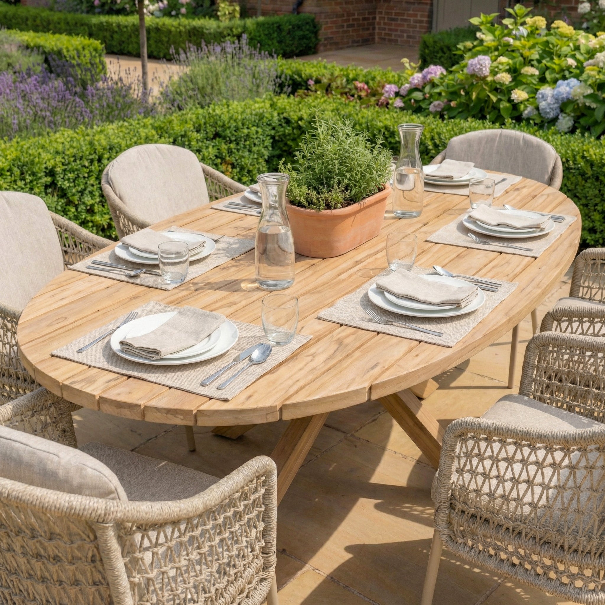 The Puccini 6 Seat Oval Dining Set in Latte features neutral tableware, woven chairs from the 4 Seasons Rope collection, and is complemented by lush garden plants.