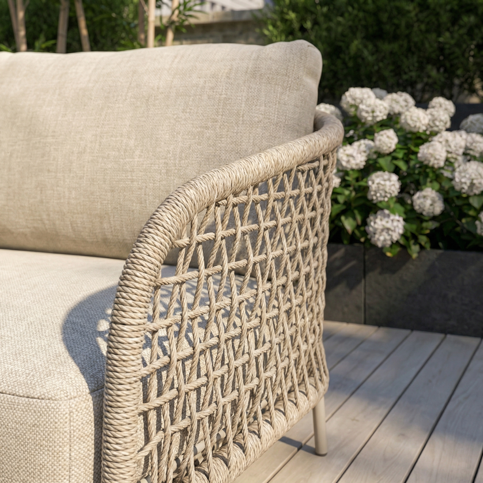Close-up of the Puccini 3 Seat Sofa (Ex Display) featuring woven rope and beige cushions on a deck, with white hydrangeas in the background.