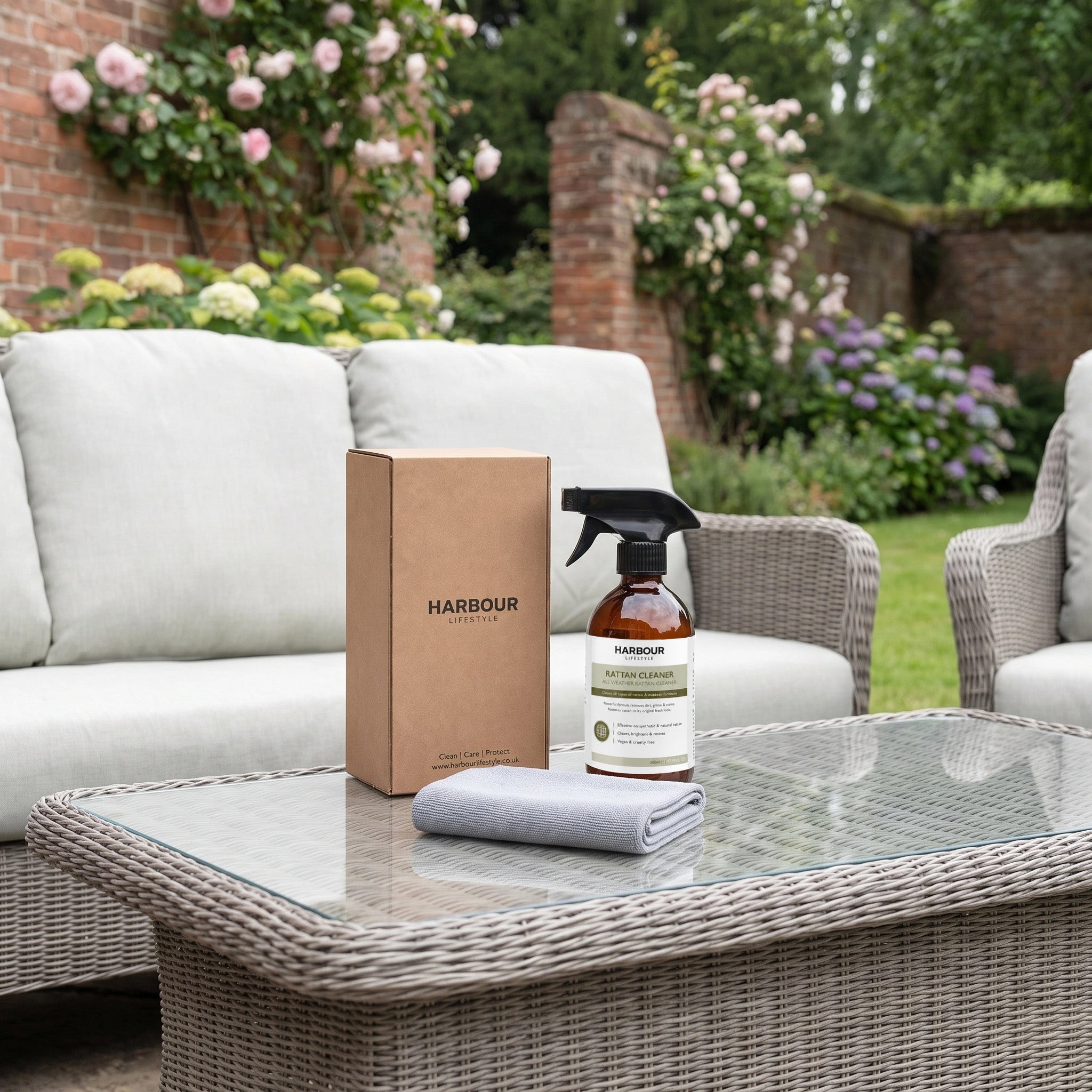 Brown box, spray bottle, and cloth on a glass table with wicker sofas and garden flowers in the background—perfect for using your Wicker and Rattan Garden Furniture Cleaner to remove dirt and grime from every surface. Ideal for maintaining rattan furniture cleaning routines.
