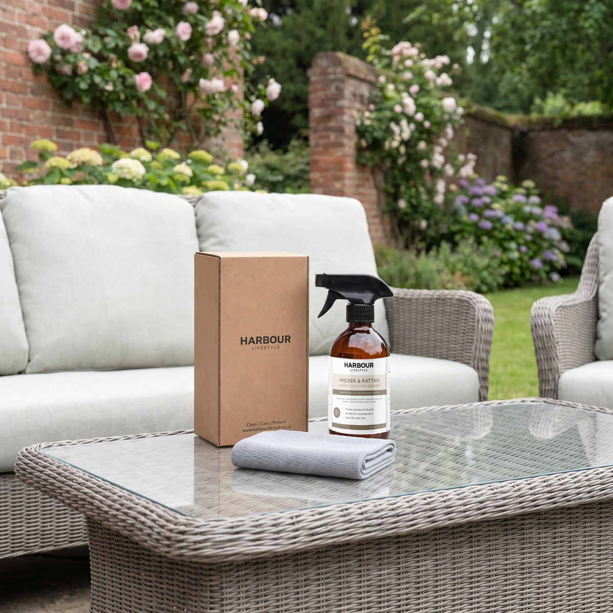 A bottle of Wicker and Rattan Garden Furniture Cleaner, along with its box and cleaning cloth, rests on a wicker outdoor table, surrounded by garden sofas and greenery—the perfect setup for removing stubborn dirt from your outdoor furniture with this effective cleaner.