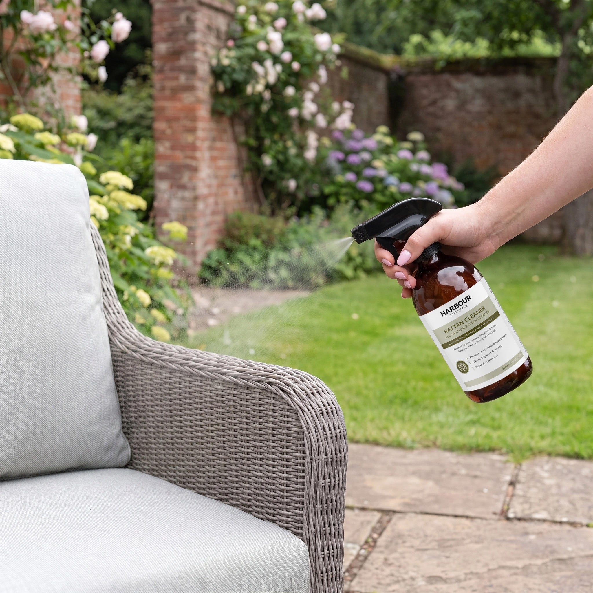 A hand sprays Wicker and Rattan Garden Furniture Cleaner onto a wicker patio chair in a garden with flowers and a brick wall, helping to remove dirt and grime for refreshed seating.