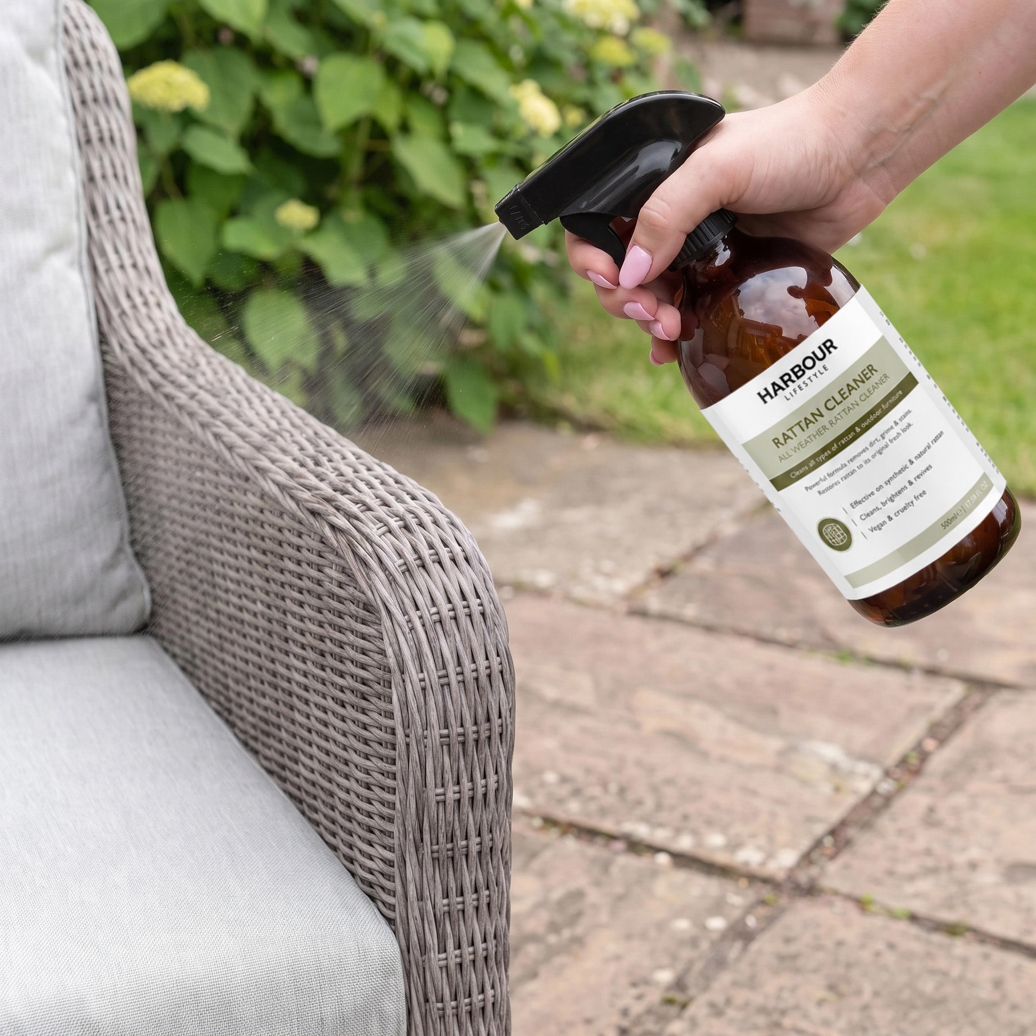 Spraying Wicker and Rattan Garden Furniture Cleaner onto a gray wicker outdoor chair, with garden plants and a stone patio in the background, to remove dirt and grime.