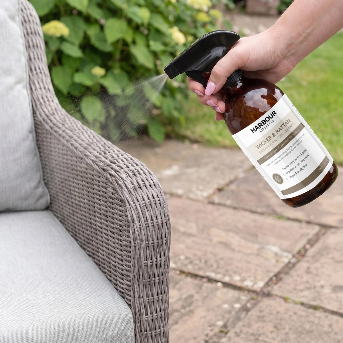 Spraying Wicker and Rattan Garden Furniture Cleaner onto a wicker patio chair outdoors on a stone patio, with plants in the background, to remove stubborn dirt and keep your seating looking fresh.