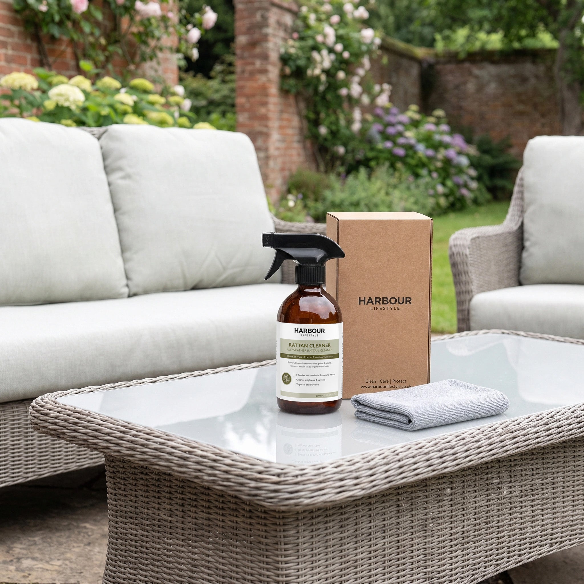 The Wicker and Rattan Garden Furniture Cleaner, along with a cleaning cloth and box, rests on a glass table between outdoor wicker sofas in a garden setting—perfect for freshening up your space and easily removing dirt and grime from rattan furniture.