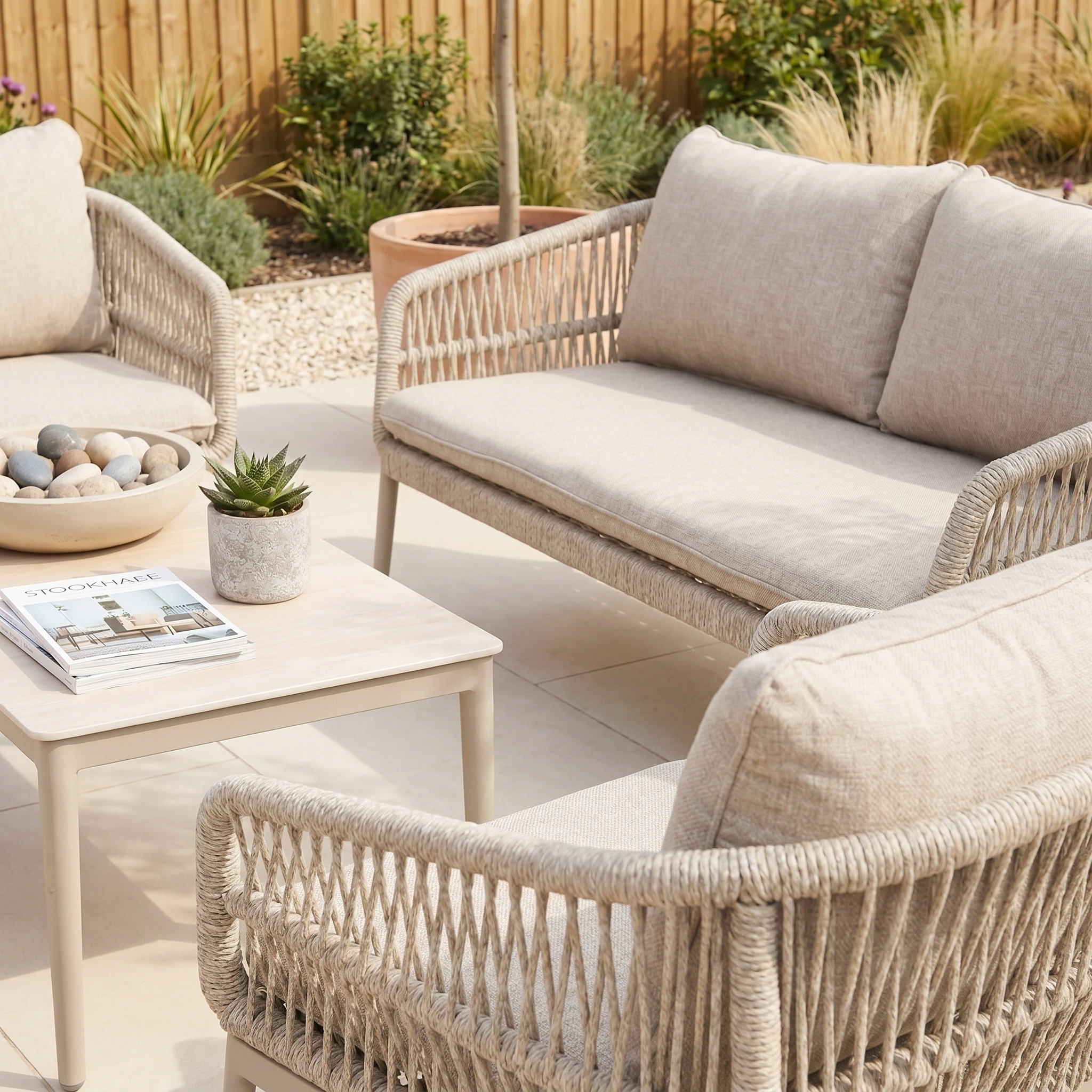 Outdoor patio with the Rhea 2 Seat Sofa Set in Latte, featuring beige rope seating, a small table, potted plant, decorative stones, and lush greenery.