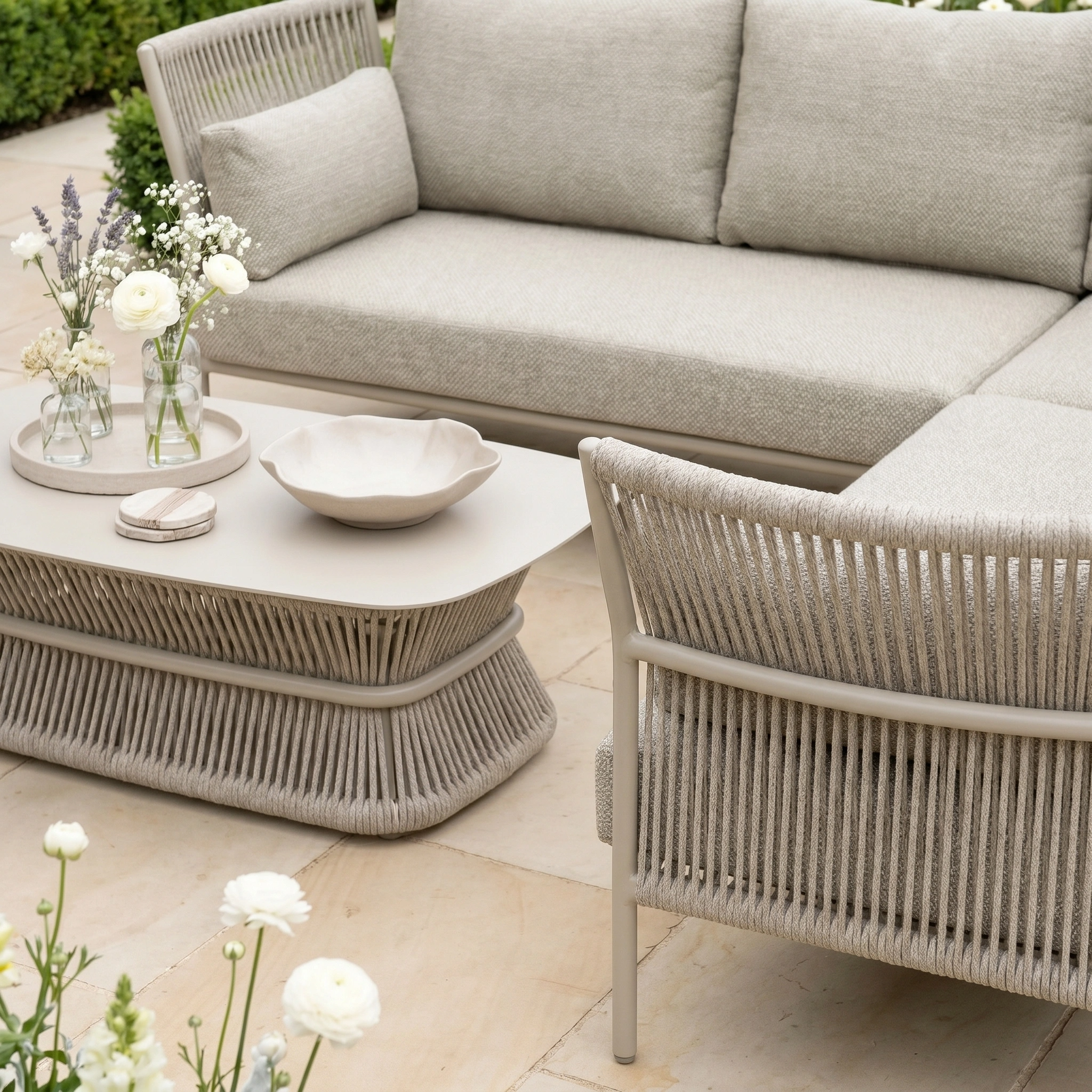 The Salina Corner Group Set with Aluminium Table in Latte features weather-resistant beige seating and a matching table, shown on a tiled patio with white flowers and a decorative bowl.