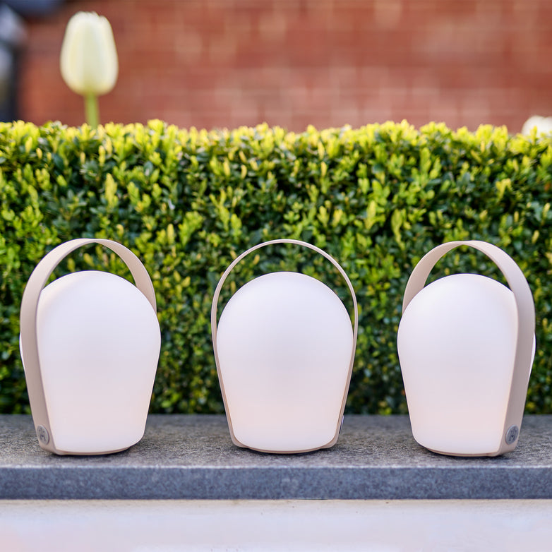 Three Bob Small Table Lights in Sand (3 Pack) are placed on a ledge in front of green shrubs, with a white flower in the background.