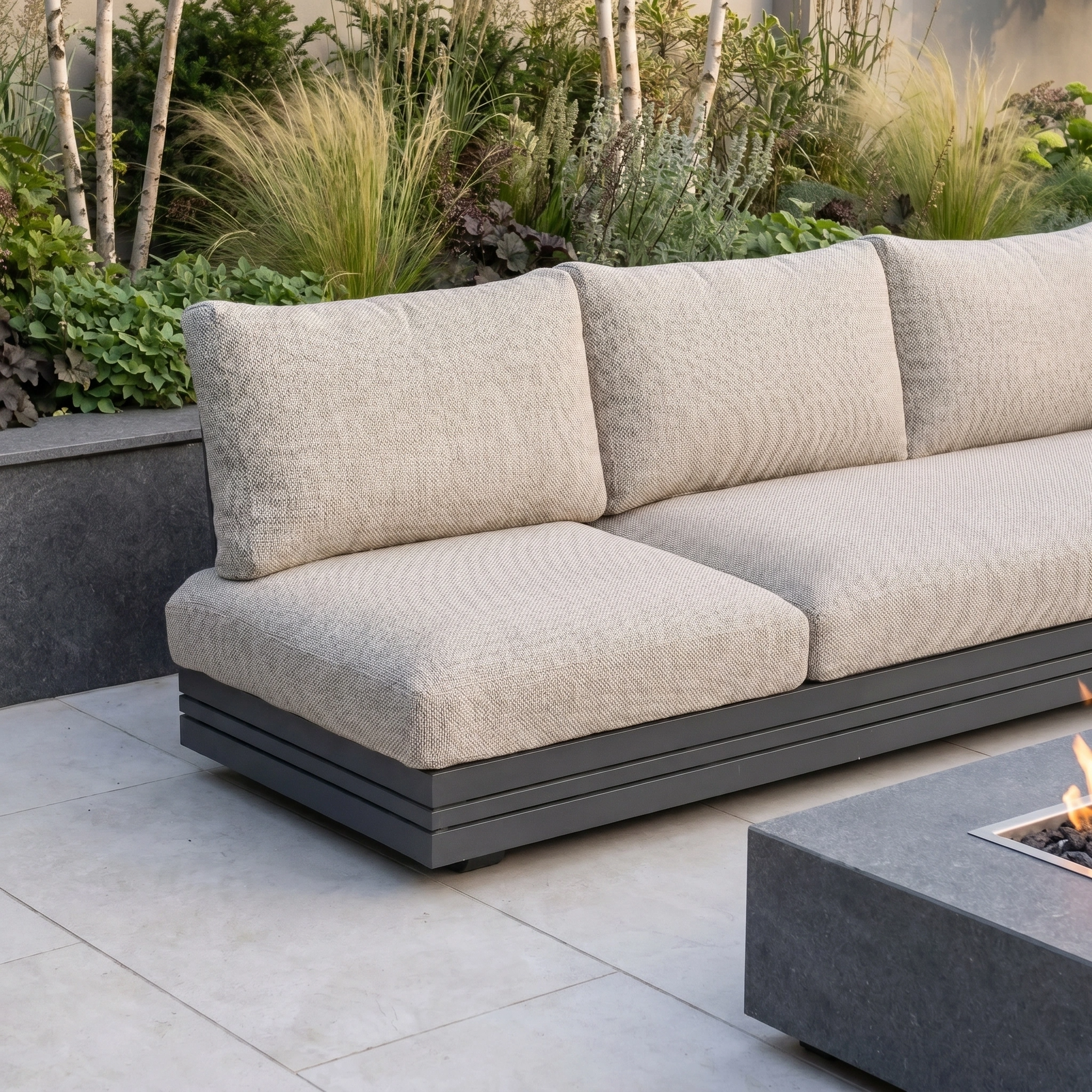 The Savona 3 Seat Sofa with Sun Lounger Feature in Truffle, designed for modern luxury, sits beside a fire pit table and lush greenery. UV-resistant, it adds comfort and style to any outdoor space.