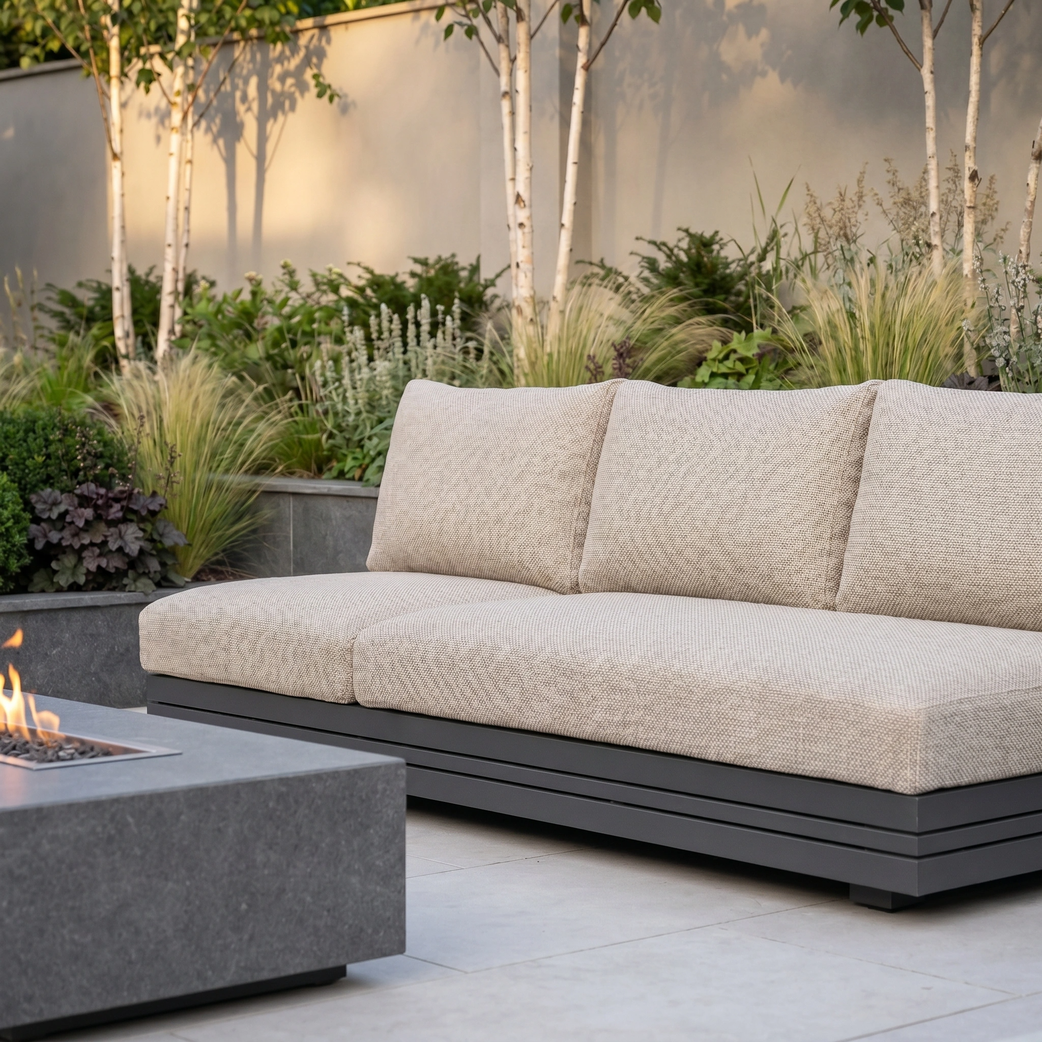 The Savona 3 Seat Sofa with Sun Lounger Feature in Truffle, a luxury UV-resistant outdoor piece, sits by a modern fire pit on the patio, surrounded by lush greenery and tall plants.