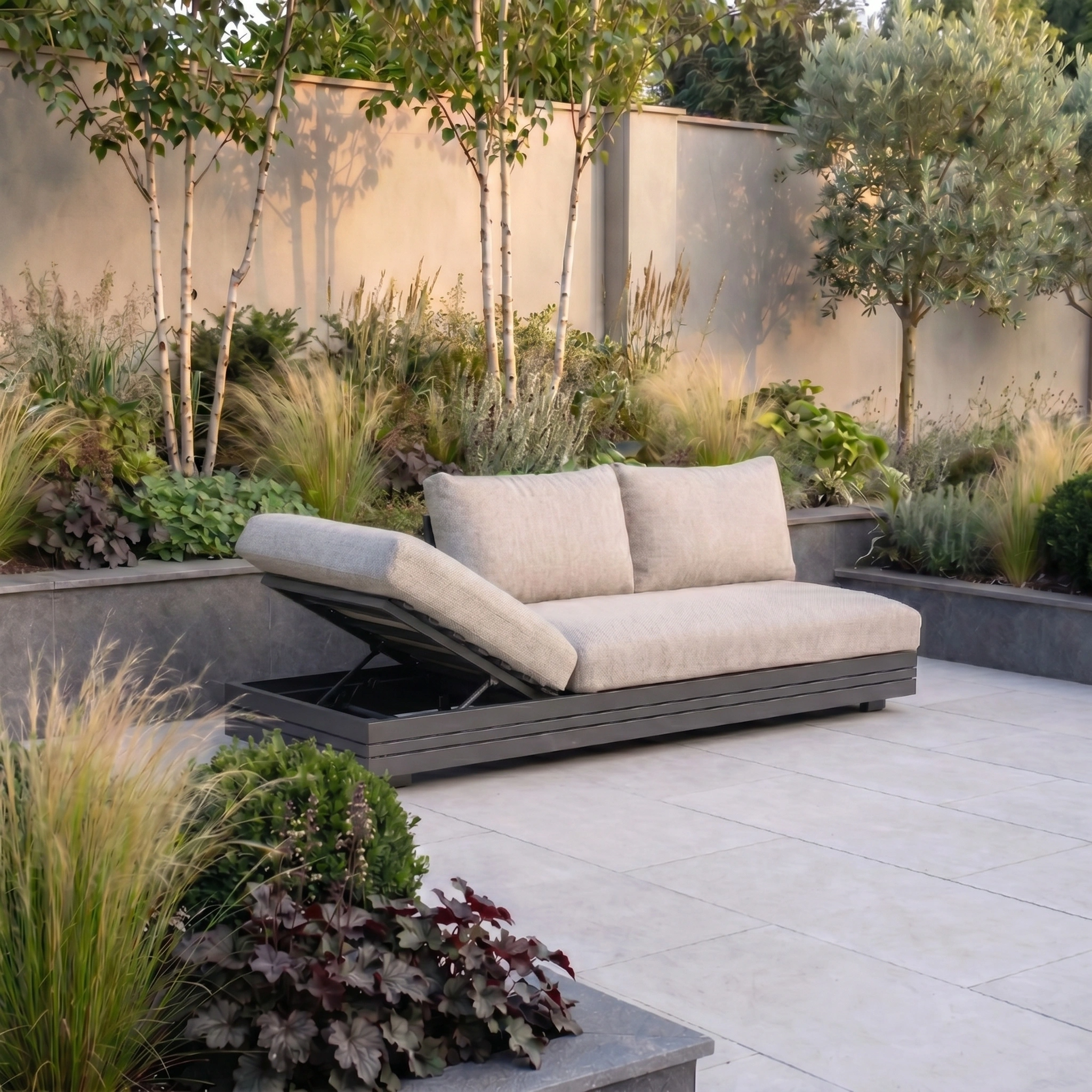 The Savona 3 Seat Sofa with Sun Lounger Feature in Truffle, designed for UV resistance, adds elegance to landscaped gardens with its beige cushions amid lush trees and ornamental grasses.