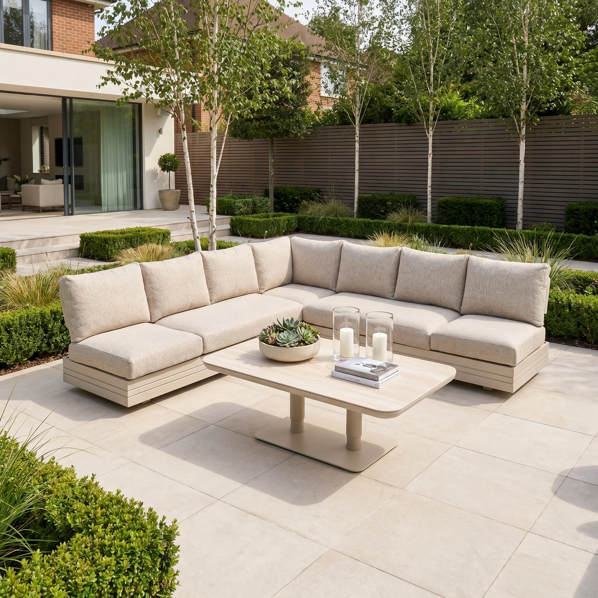 The Savona Luxury Outdoor Large Corner Sofa Set with Altura Rising Table in Oatmeal offers elegant, modern comfort on a tiled patio amid lush landscaping—a modular garden furniture set perfect for stylish outdoor living.