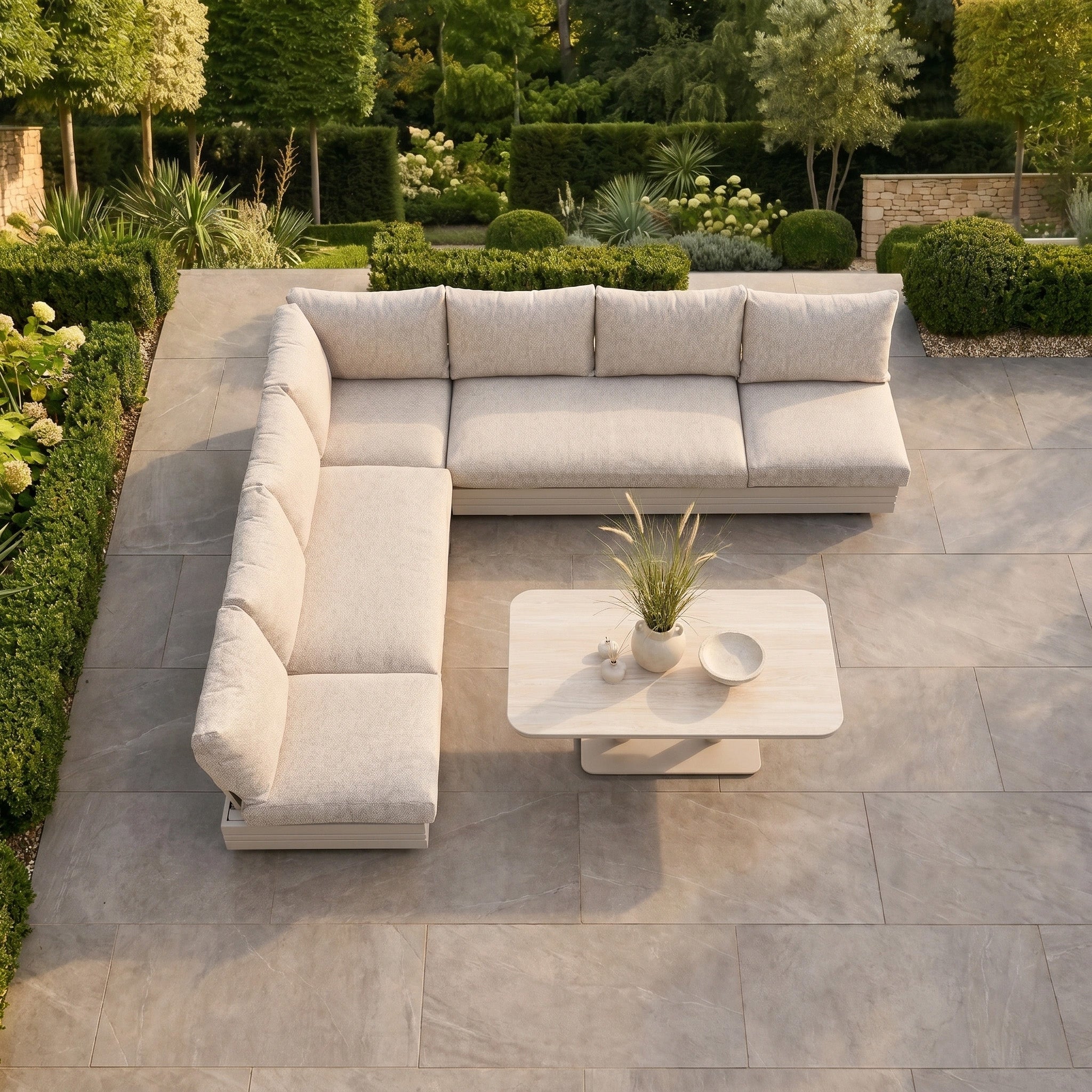 The Savona Luxury Outdoor Large Corner Sofa Set with Altura Rising Table in Oatmeal is displayed on a tiled patio with plants, surrounded by lush green garden landscaping.