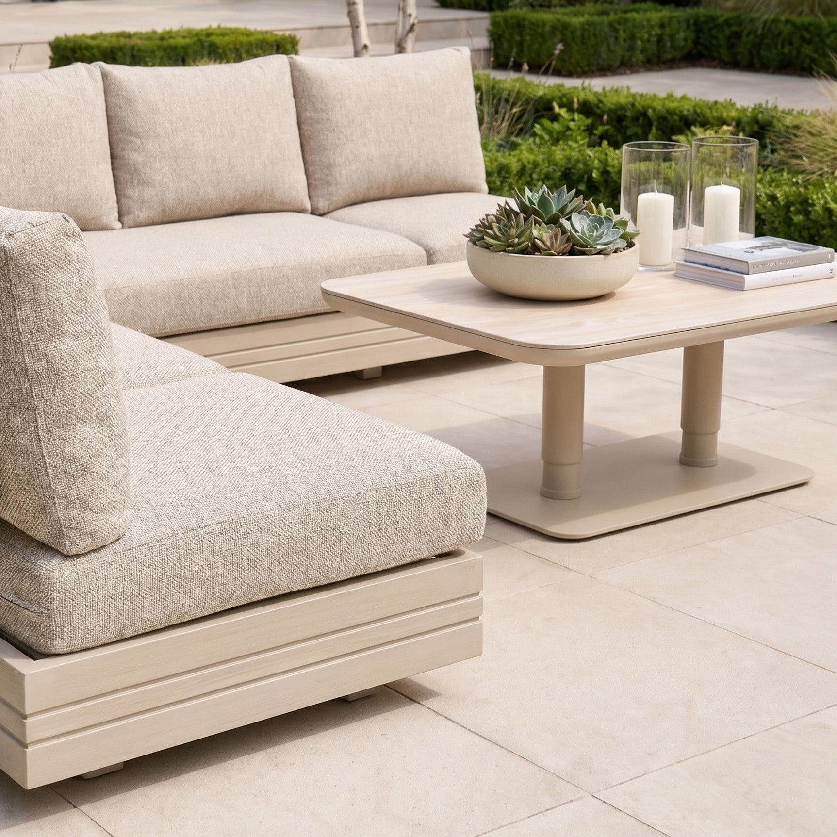 The Savona Luxury Outdoor Large Corner Sofa Set with Altura Rising Table in Oatmeal features plush seating and a versatile table, creating an elegant outdoor retreat perfect for relaxing with candles, a book, and a succulent centerpiece.