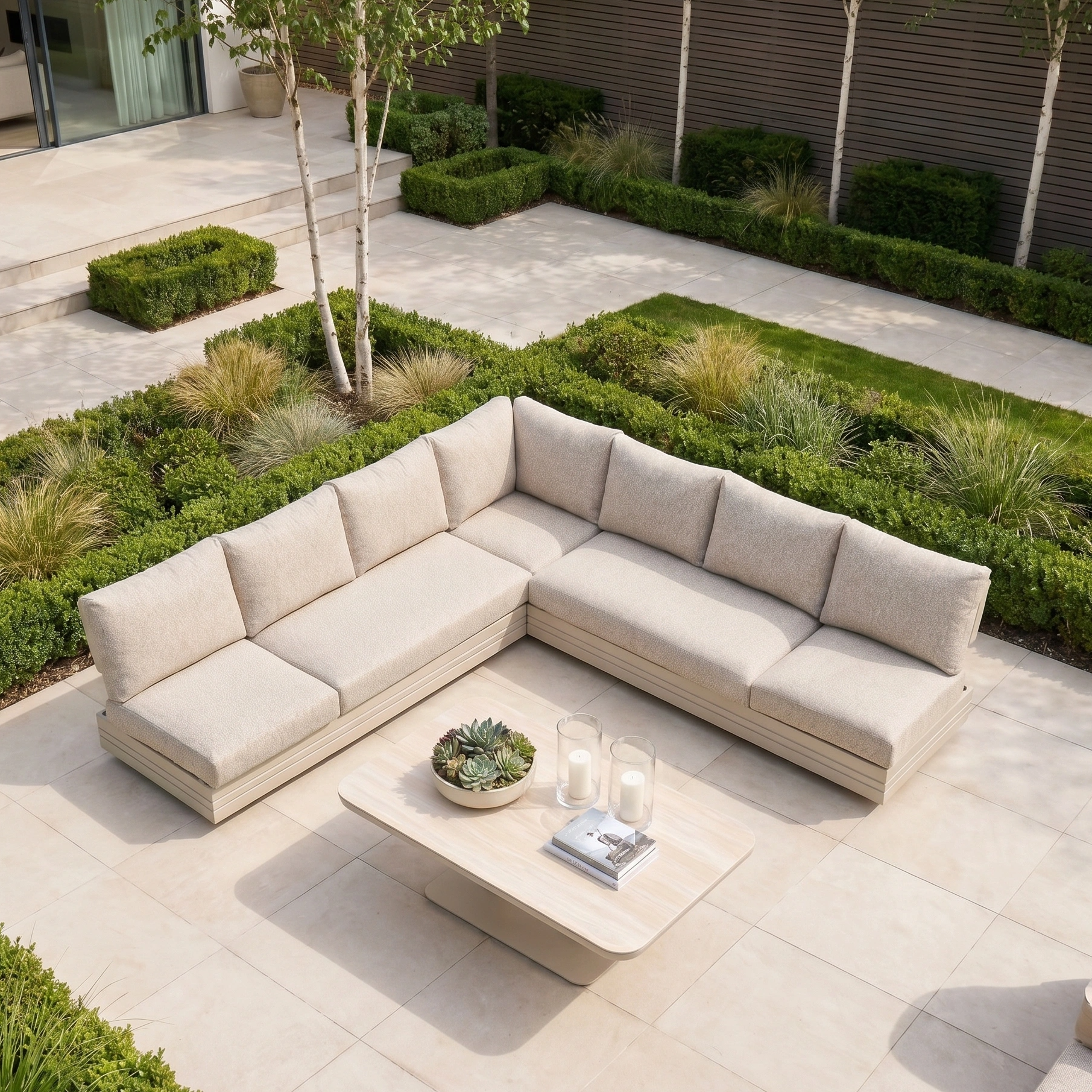 The Savona Luxury Outdoor Large Corner Sofa Set with Altura Rising Table in Oatmeal is styled with decor on a light patio surrounded by lush greenery, creating an inviting and elegant garden retreat.
