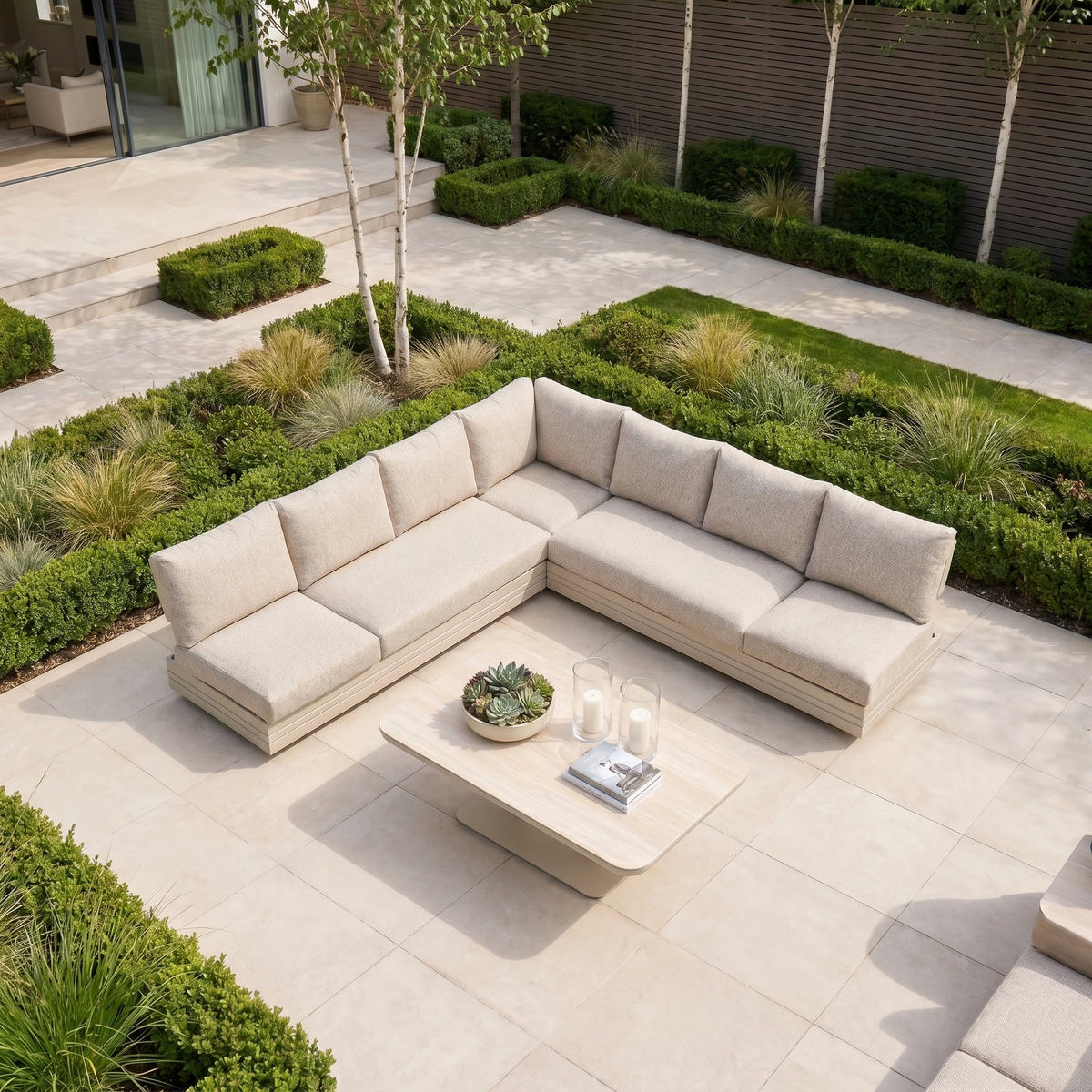The Savona Luxury Outdoor Large Corner Sofa Set with Altura Rising Table in Oatmeal is styled with decor on a light patio surrounded by lush greenery, creating an inviting and elegant garden retreat.