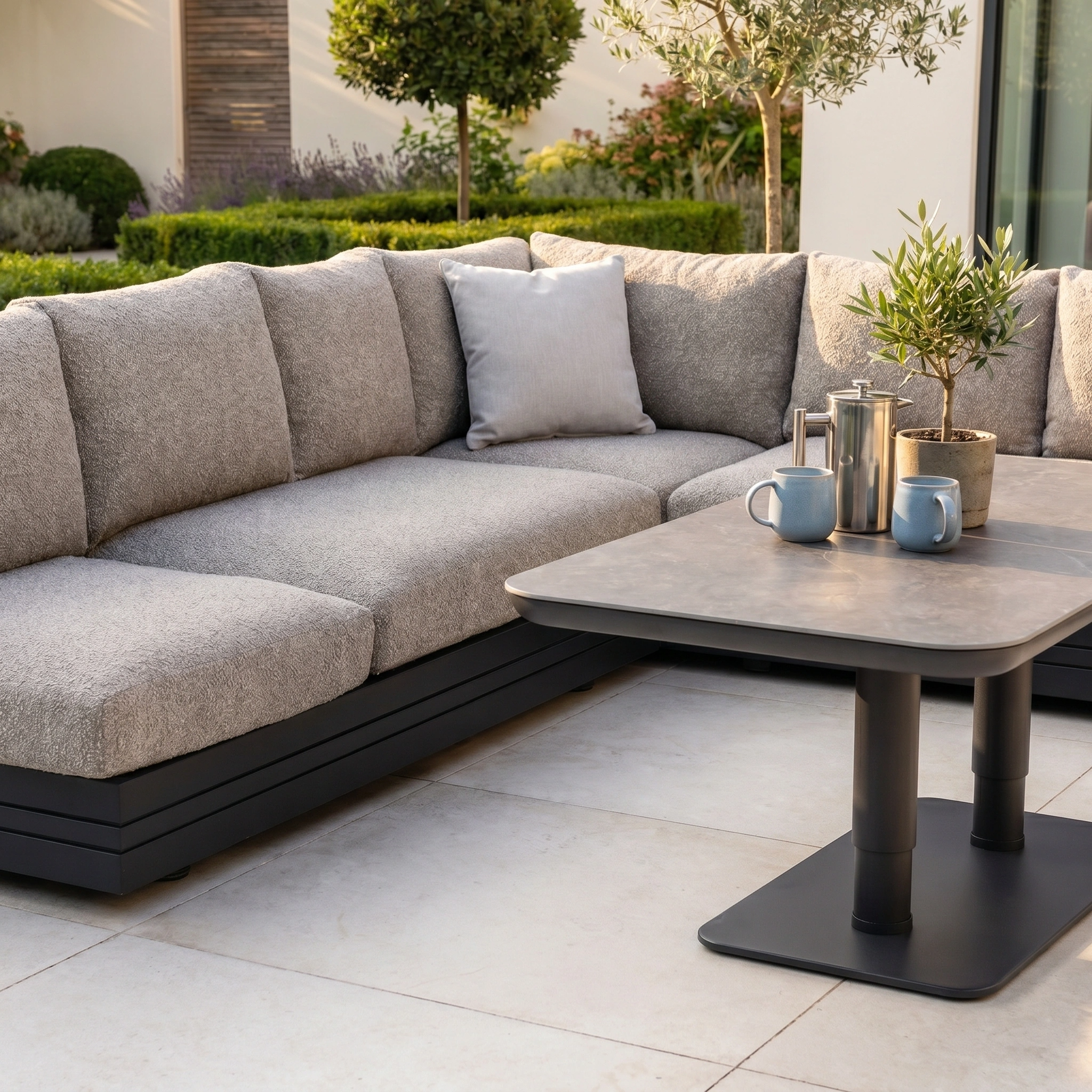 Modern outdoor patio featuring the Savona Luxury Outdoor Large Corner Sofa Set with Altura Rising Table in Truffle, paired with potted plants and metal drinkware on a tiled floor.