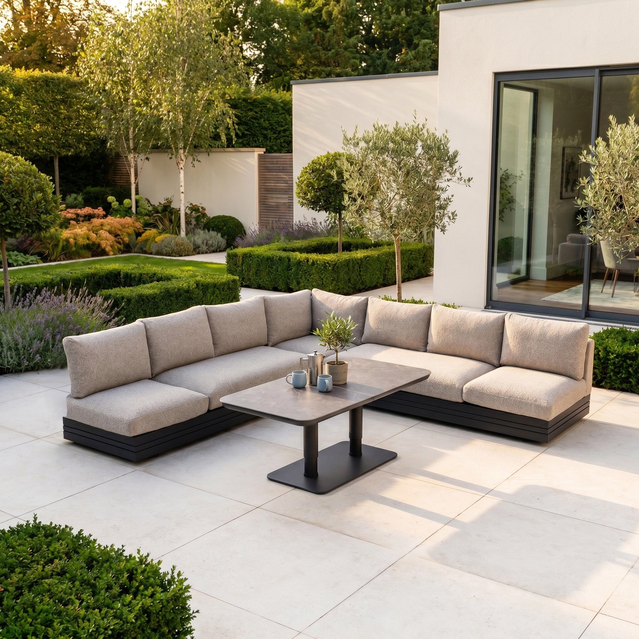 Enjoy a modern outdoor patio with the Savona Luxury Outdoor Large Corner Sofa Set in Truffle, featuring an Altura rising table, perfect for relaxing in lush green garden surroundings.