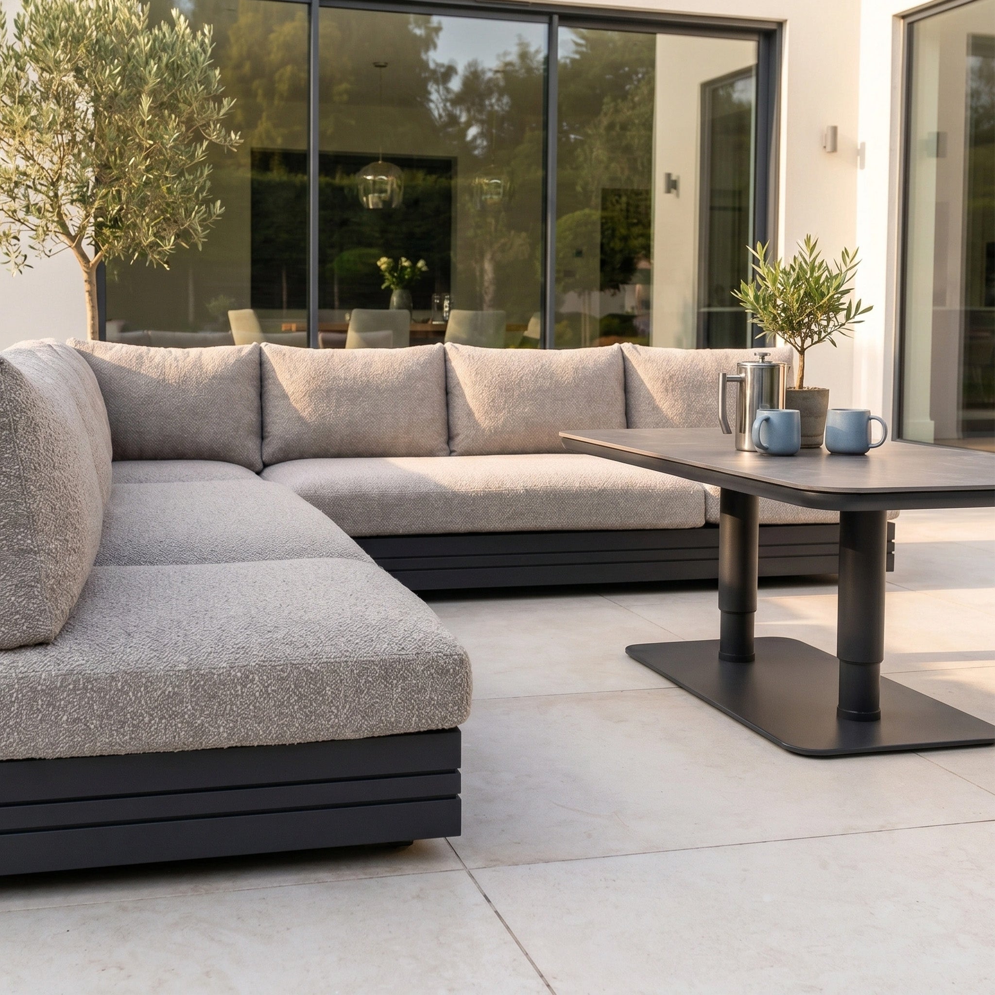 Contemporary patio with the Savona Luxury Outdoor Large Corner Sofa Set in Truffle and Altura Rising Table, complemented by lush plants and mugs beside sleek glass doors.