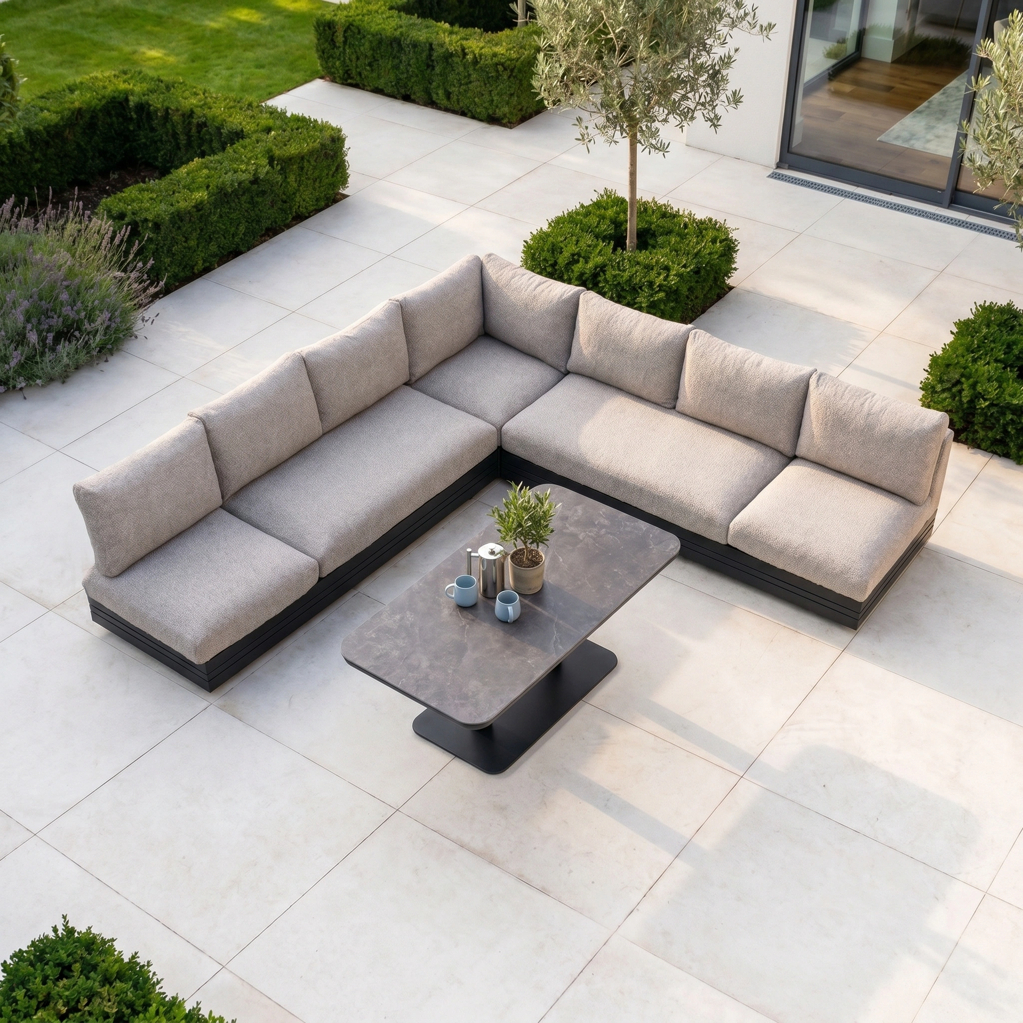 Outdoor patio featuring the Savona Luxury Outdoor Large Corner Sofa Set with Altura Rising Table in Truffle on a tiled terrace, surrounded by greenery.