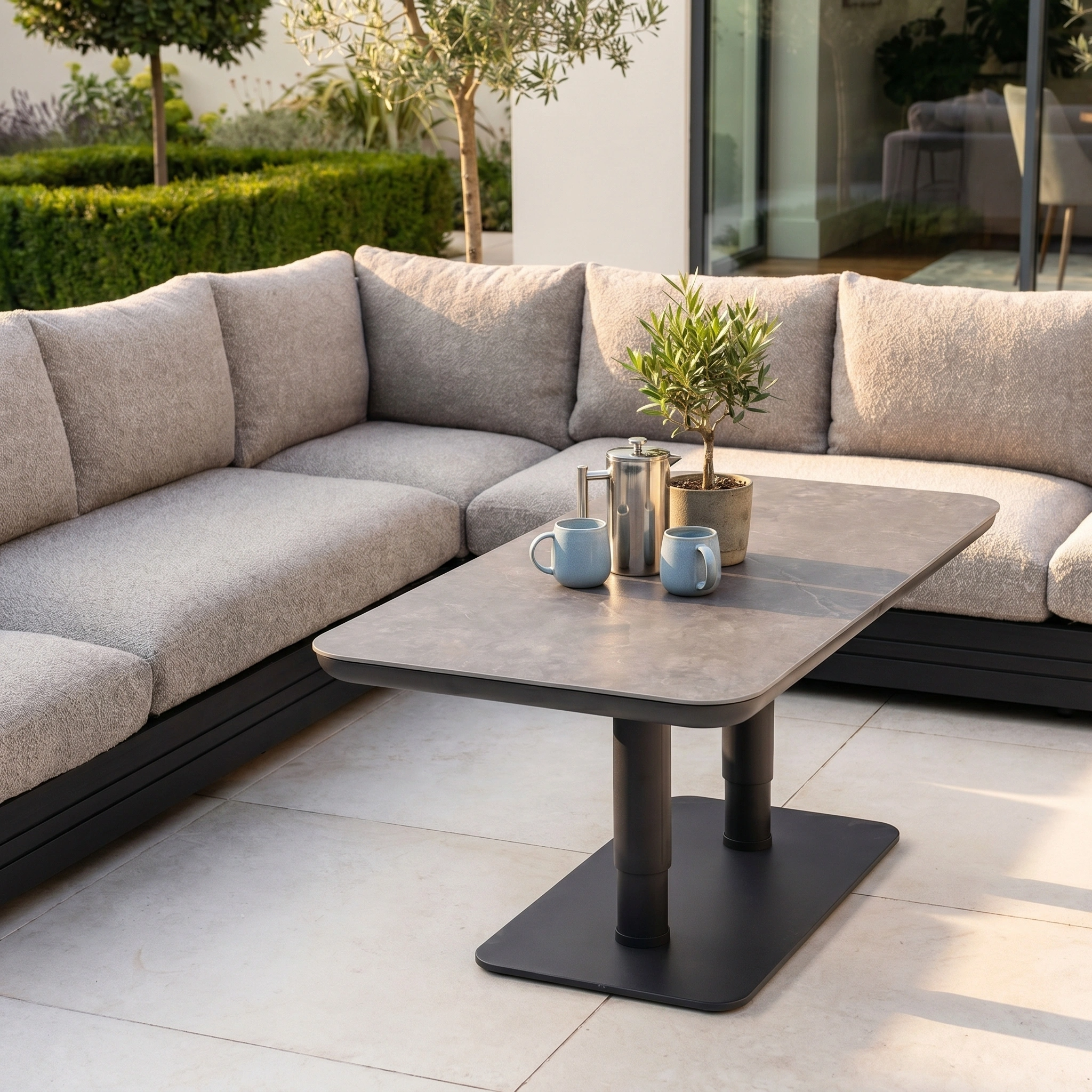 Outdoor patio with the Savona Luxury Outdoor Large Corner Sofa Set and Altura Rising Table in Truffle, mugs, teapot, and lush potted plants on a tiled floor.