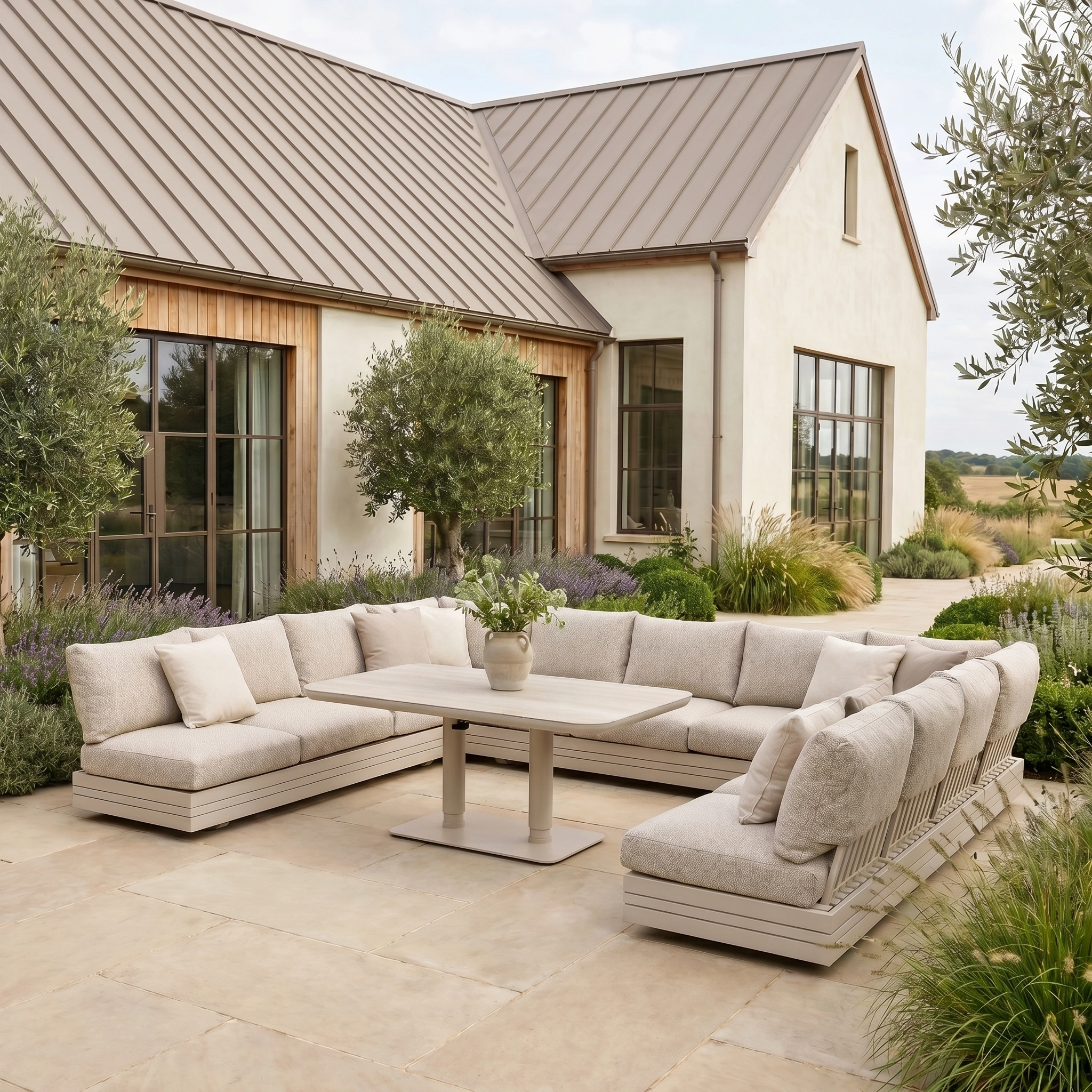 The Savona Luxury Outdoor Large U-Shape Corner Sofa Set with Altura Rising Table in Oatmeal sits on a stone patio beside a house with large windows and lush greenery.